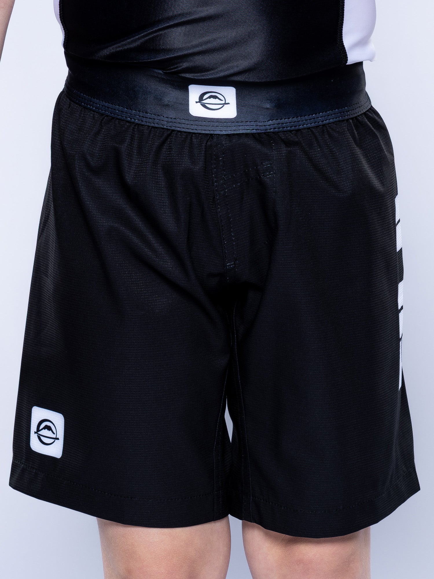 A person wearing Baseline Grappling Kids Shorts—black with white logo patches on the waistband and left leg—stands against a plain light background.