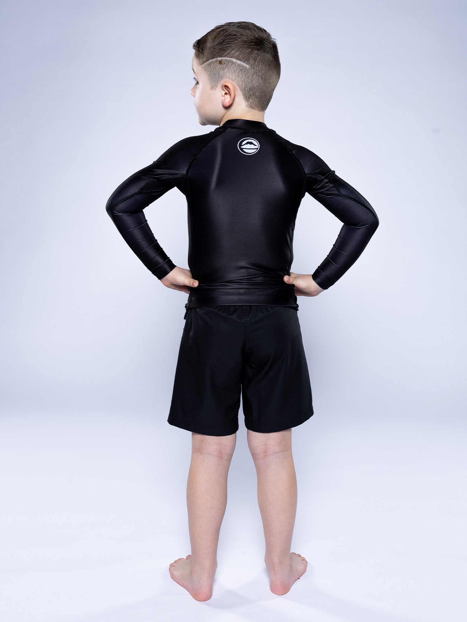 A young boy stands barefoot on a white background, facing away with hands on hips, wearing the Kids Baseline 2.0 Long Sleeve Rashguard Black and black shorts.