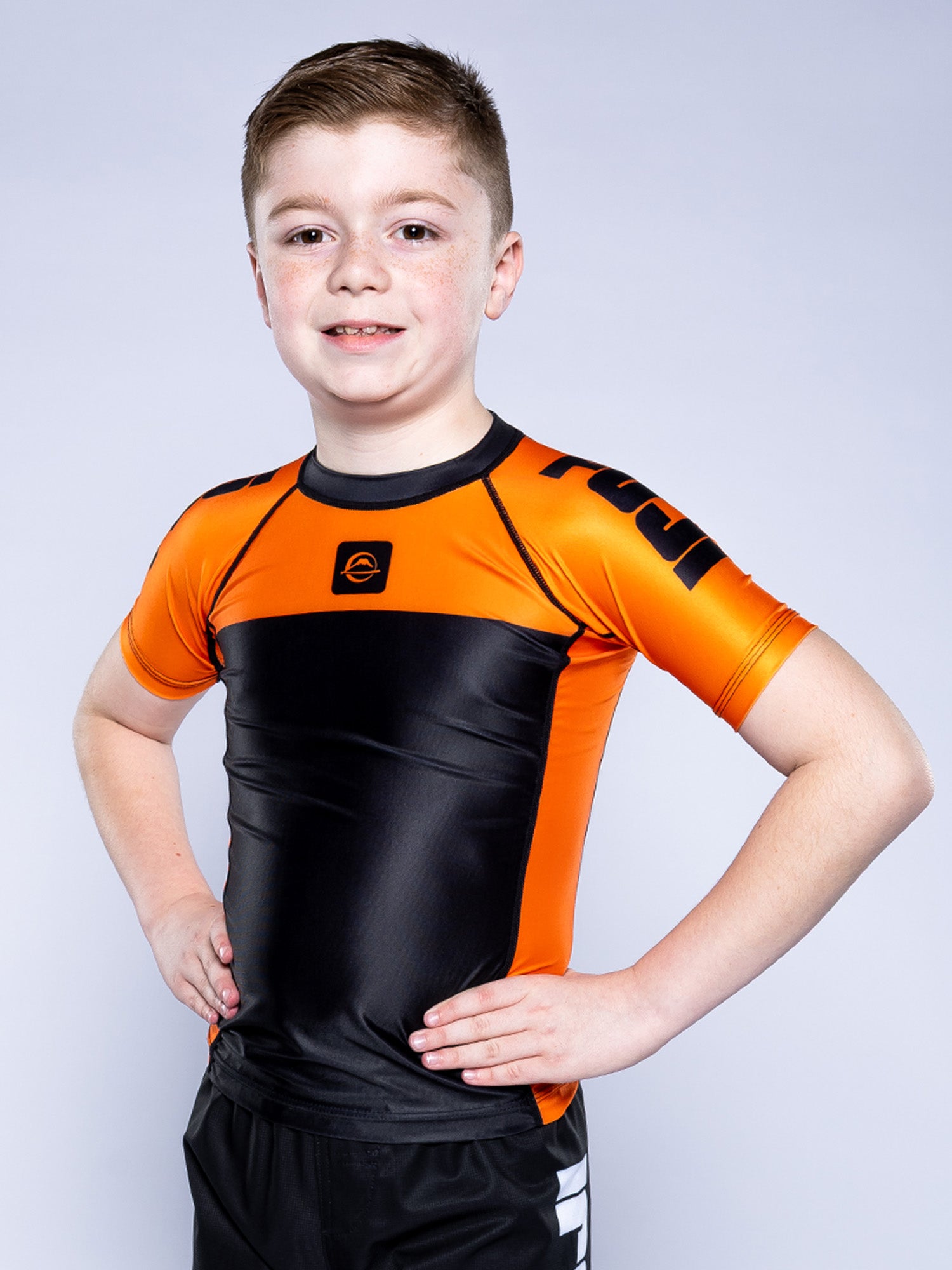 A young boy with short light brown hair smiles confidently, wearing the Kids Competition Ranked Rashguard Orange and black shorts, standing with his hands on his hips against a light gray background.