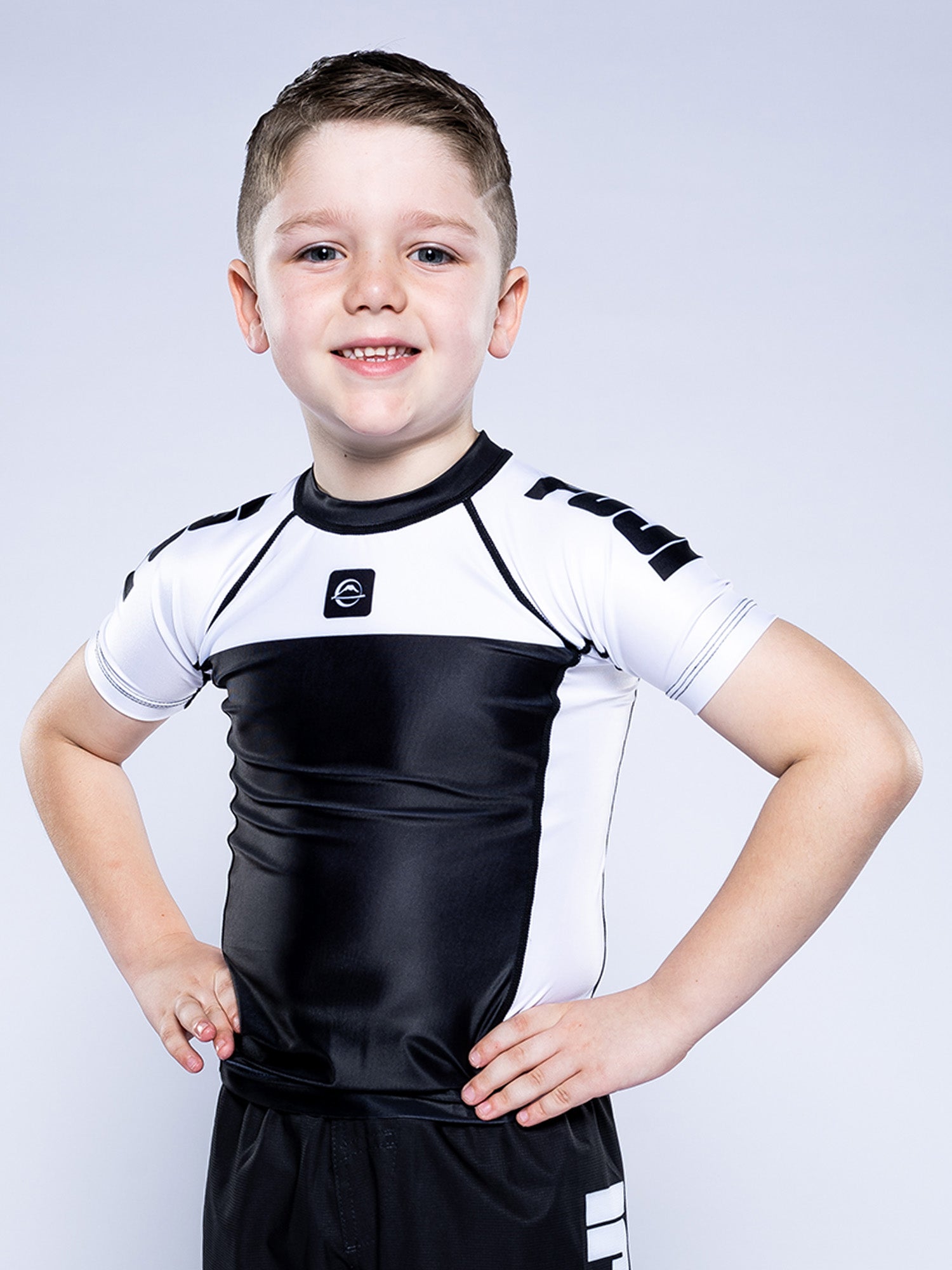 A young boy with short brown hair stands confidently with hands on hips, wearing the Kids Competition Ranked Rashguard White and black shorts, posed against a plain light gray background.