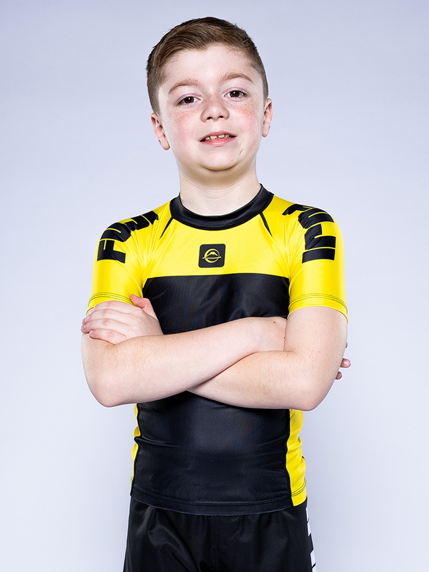 A young boy with short brown hair stands with arms crossed, smiling, wearing the Kids Competition Ranked Rashguard Yellow and black shorts against a plain light gray background.