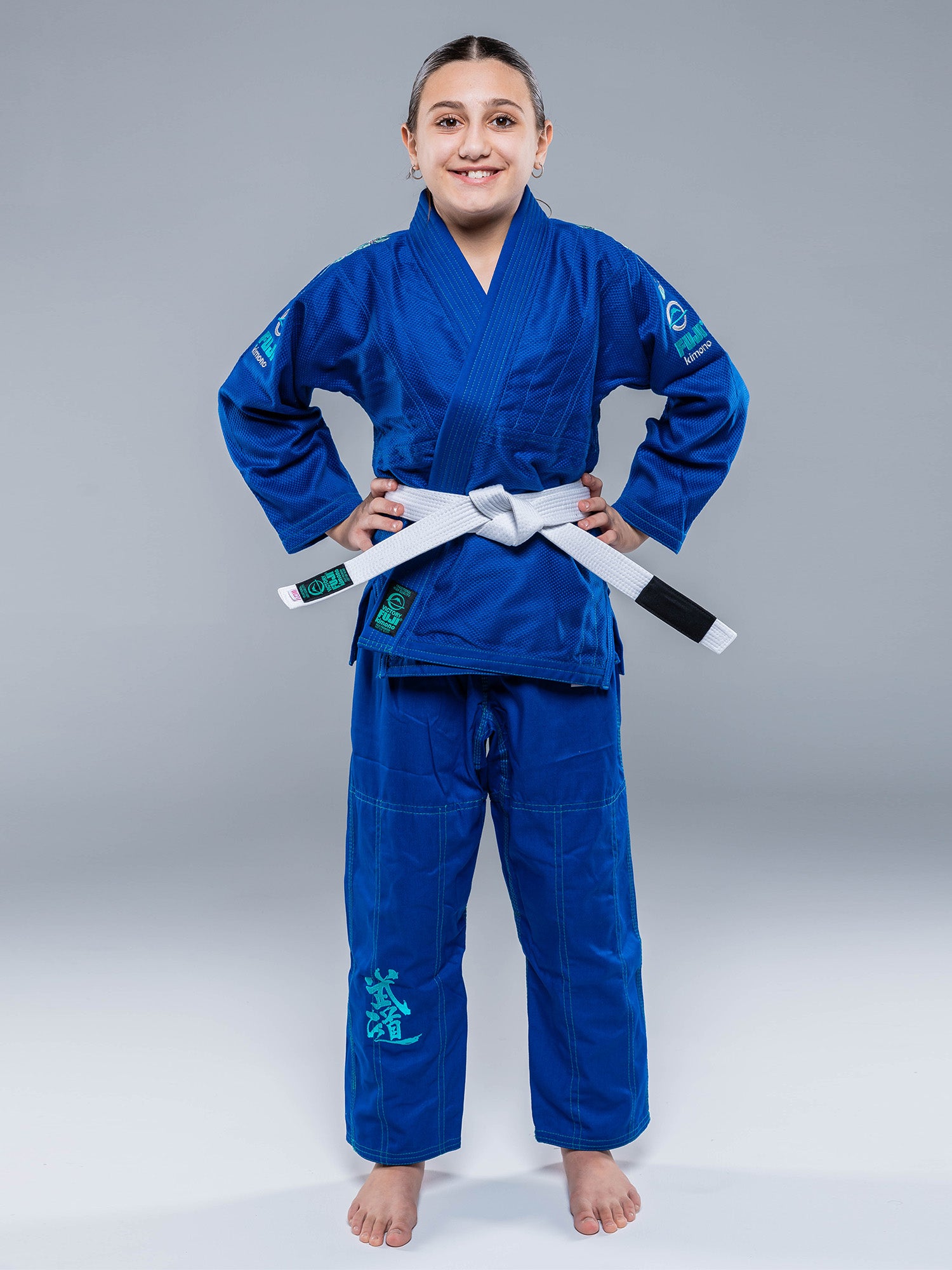 A young person smiles with hands on hips, barefoot on a white surface, wearing the Blossom Kids Jiu Jitsu Gi Blue and a white belt, against a plain gray background.