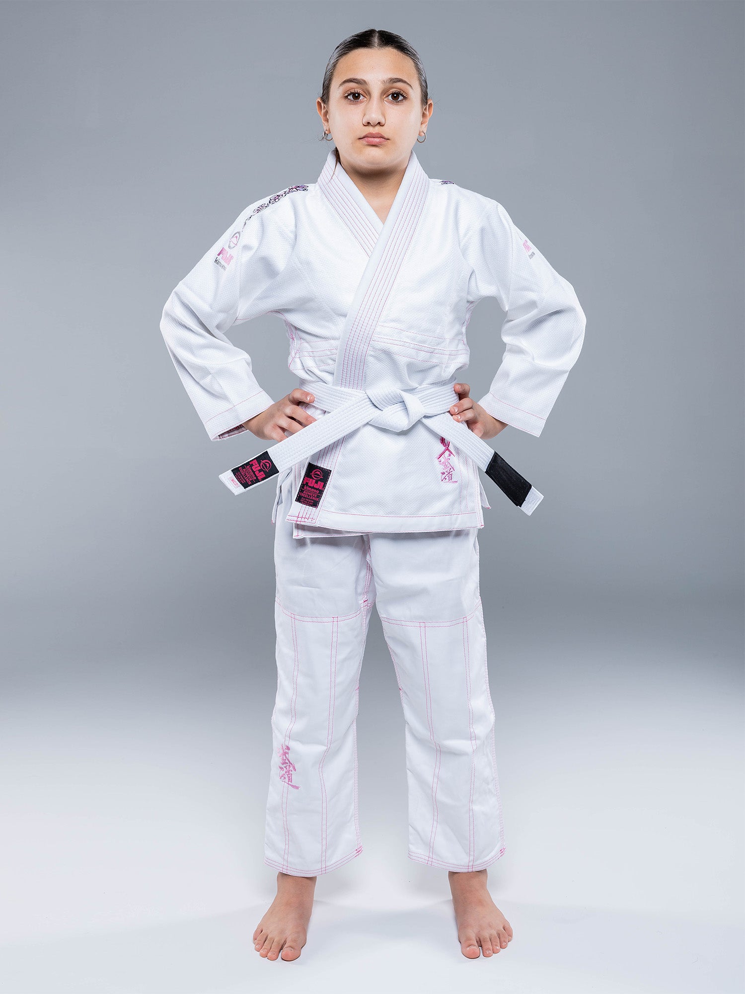 A young person, barefoot, wears the Blossom Kids Jiu Jitsu Gi White with a white belt and stands facing forward with a serious look against a plain gray background.