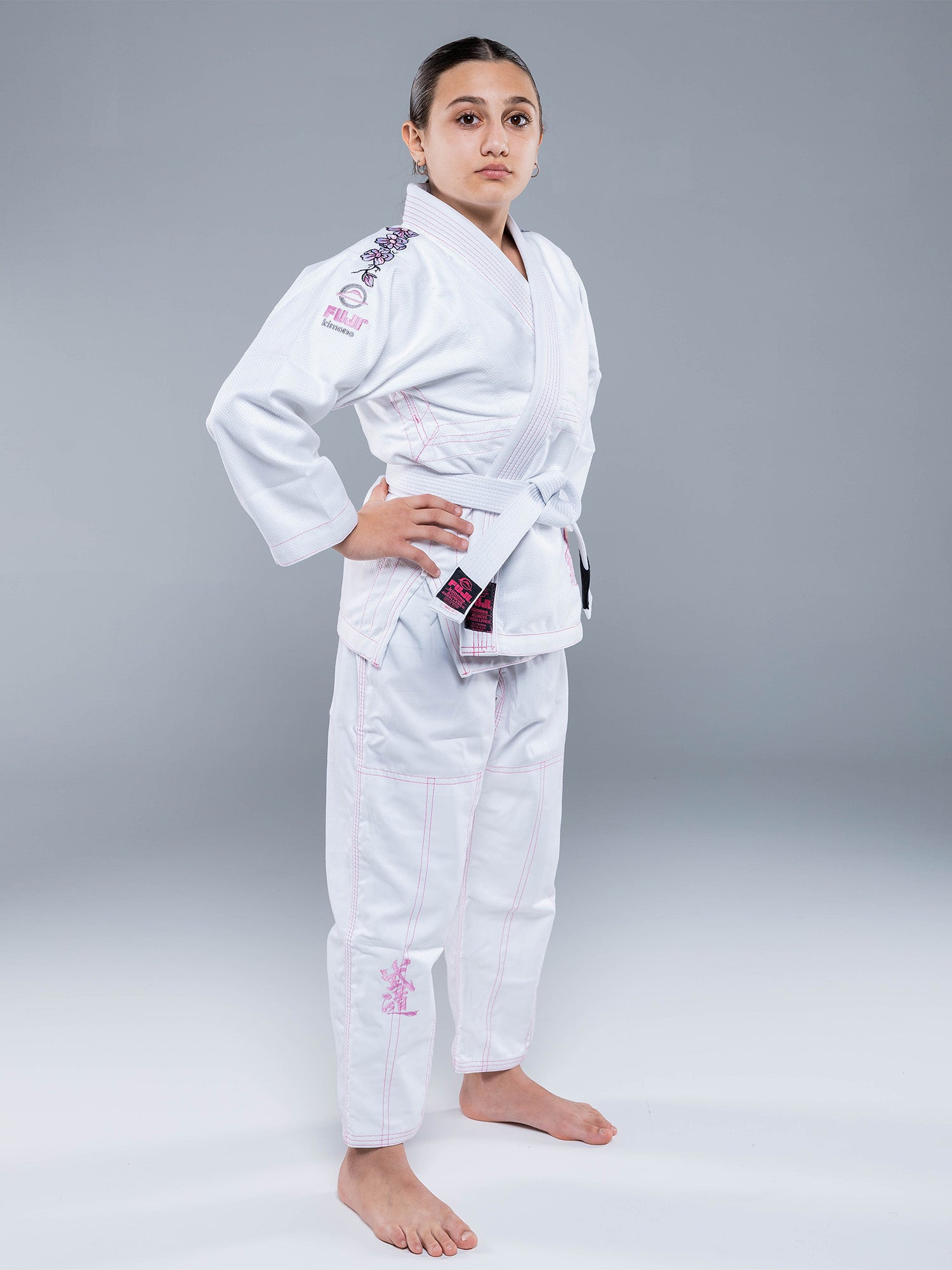 A young person stands confidently in the Blossom Kids Jiu Jitsu Gi White, featuring pink stitching and a white belt, with bare feet against a plain grey background. Their hands rest on their hips.