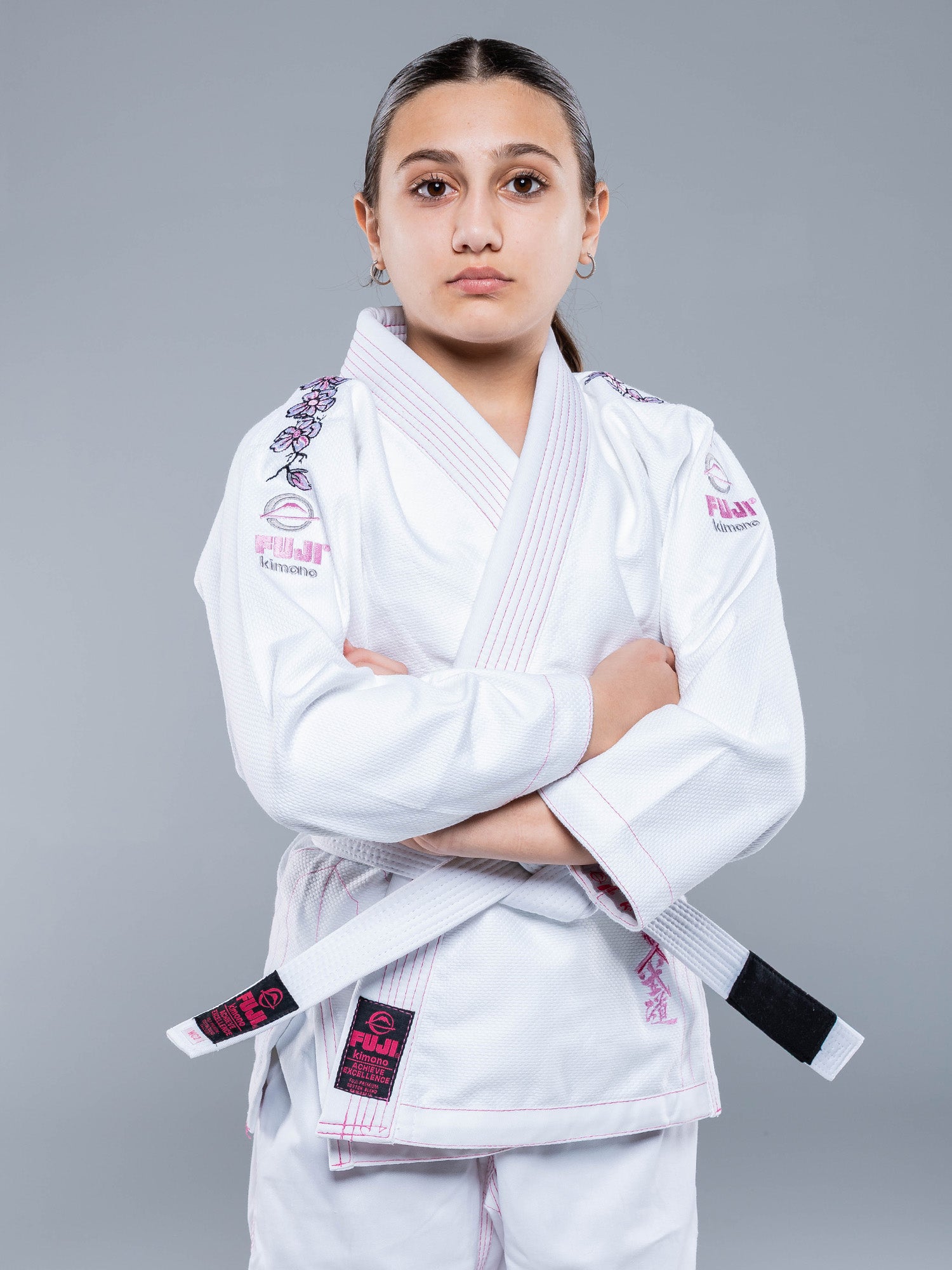 A young girl stands confidently with arms crossed, wearing the Blossom Kids Jiu Jitsu Gi White with pink accents and a white belt, set against a plain gray background.