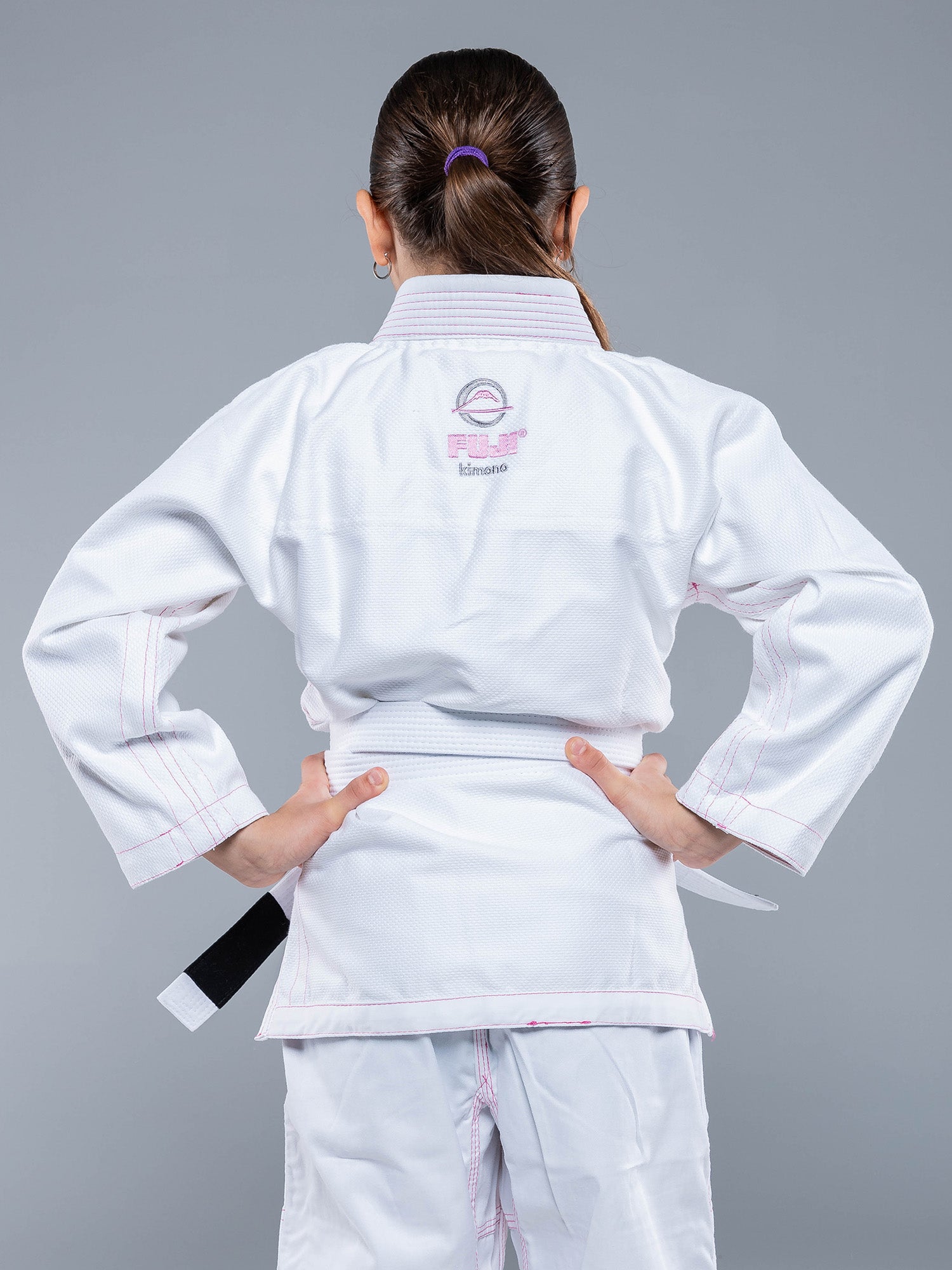 A child stands with hands on hips, wearing the Blossom Kids Jiu Jitsu Gi White and a white belt, facing away against a plain gray background.