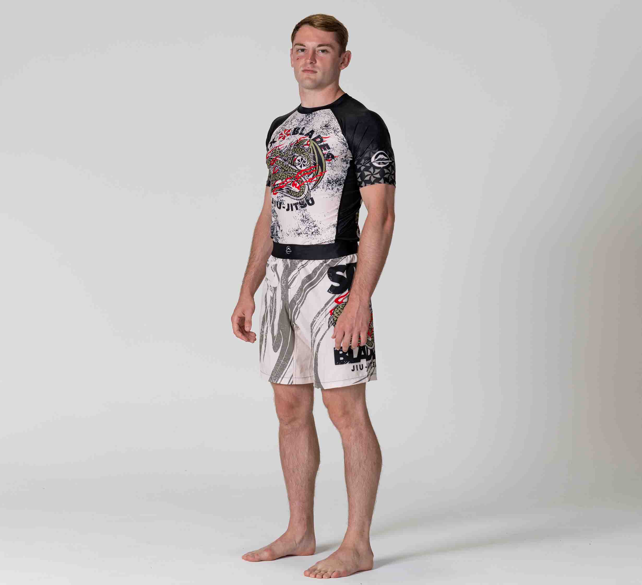 A young man stands barefoot in a studio, wearing Six Blades Dragon Flex Lite Shorts with matching black and white rash guard featuring red, black, and gray graphics including "BJJ" and "Jiu Jitsu," facing slightly to the side against a plain background.