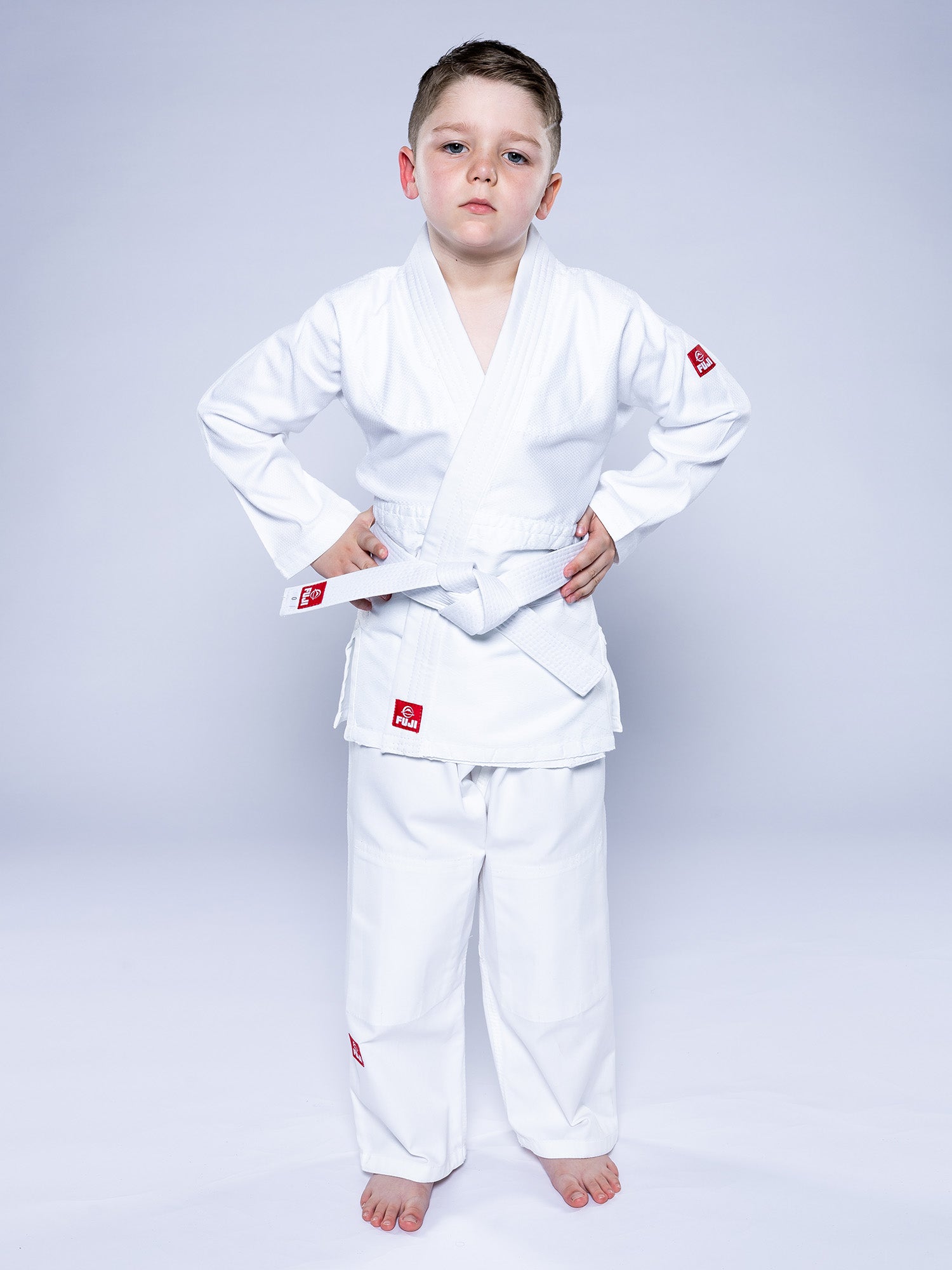 A young boy stands barefoot, hands on hips, confidently modeling the Essential Judo Kimono White against a plain light background.