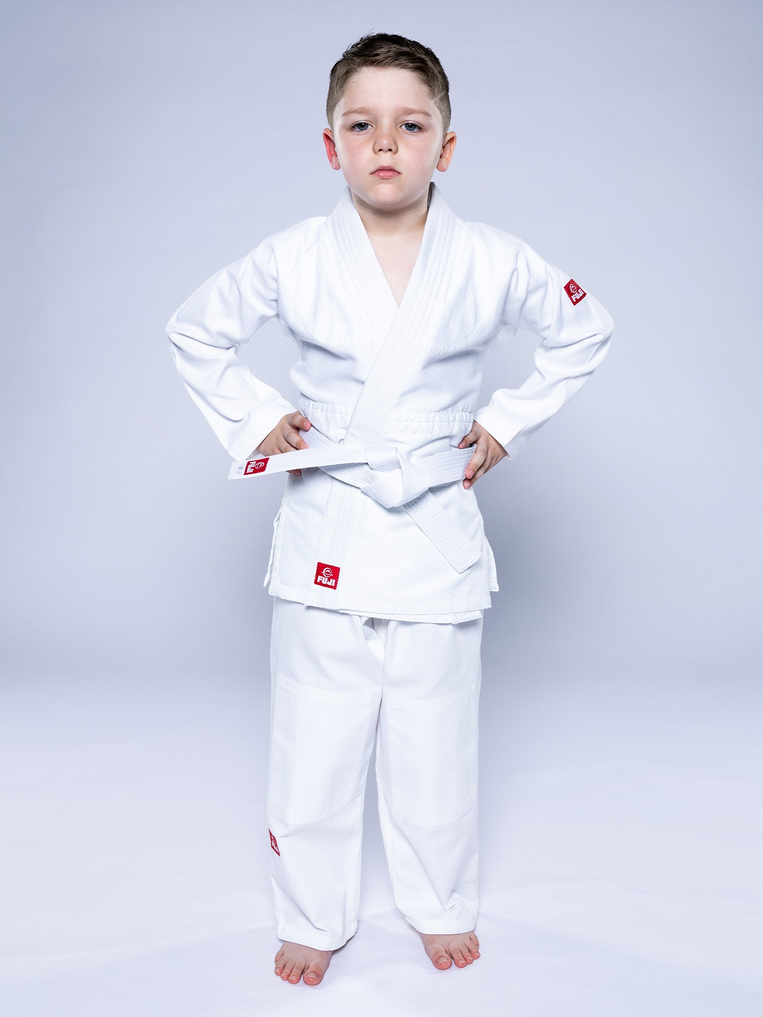 A young boy stands barefoot on a white surface, hands on hips with a serious expression, wearing the Essential Judo Kimono White and belt against a plain light gray background.