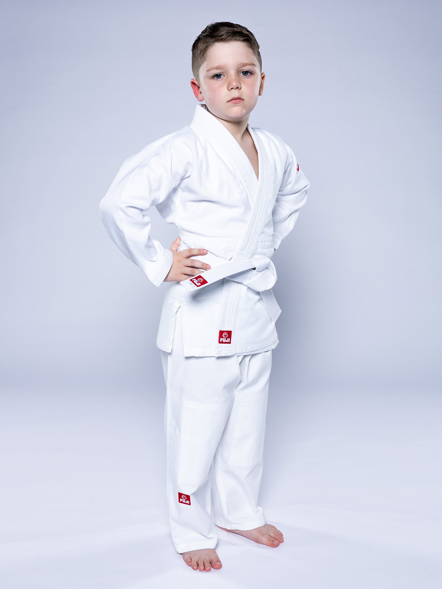 A young boy stands barefoot against a plain light background, hands on his hips and wearing the Essential Judo Kimono White, with a serious expression on his face.