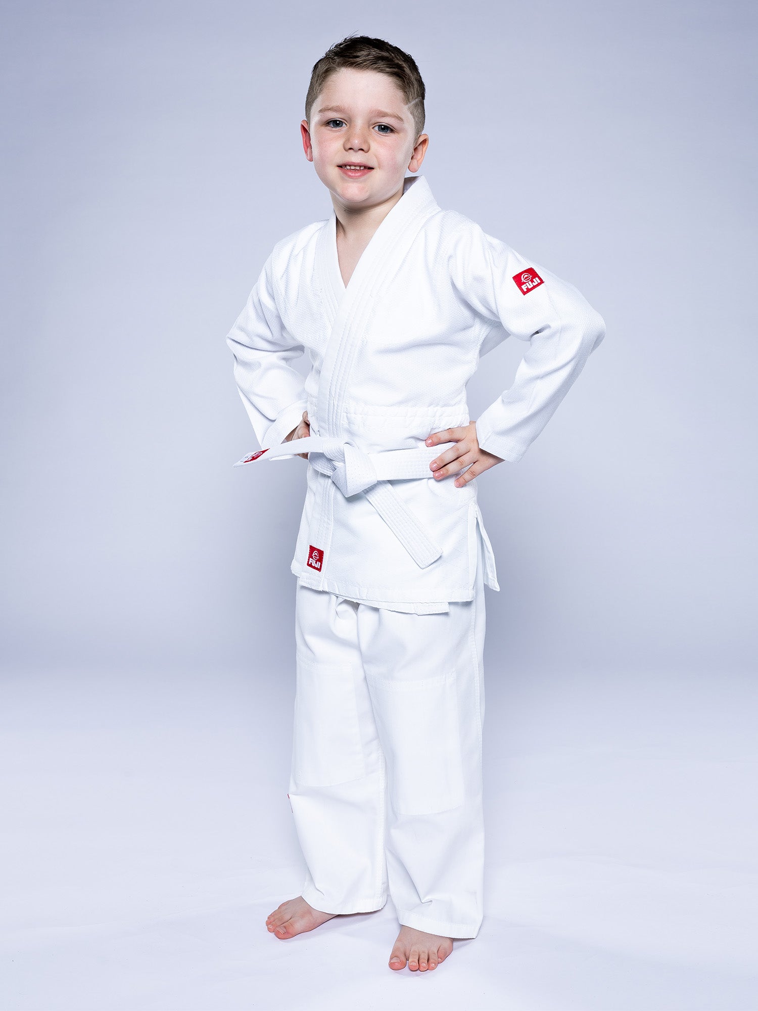 A young boy, barefoot and smiling with hands on hips, wears the Essential Judo Kimono White martial arts uniform against a light background.