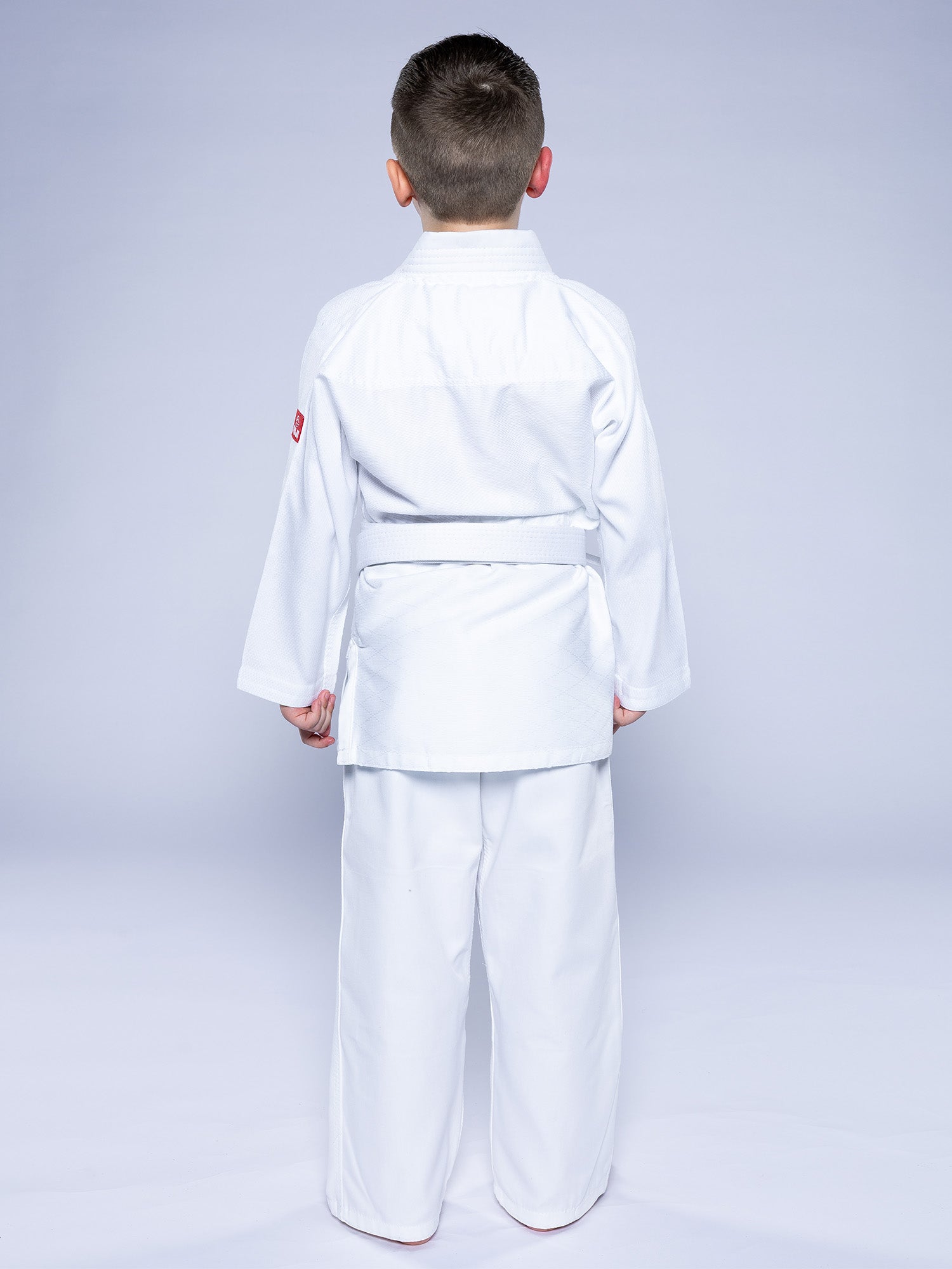 A young boy faces away from the camera, dressed in the Essential Judo Kimono White with a white belt, set against a plain light background.