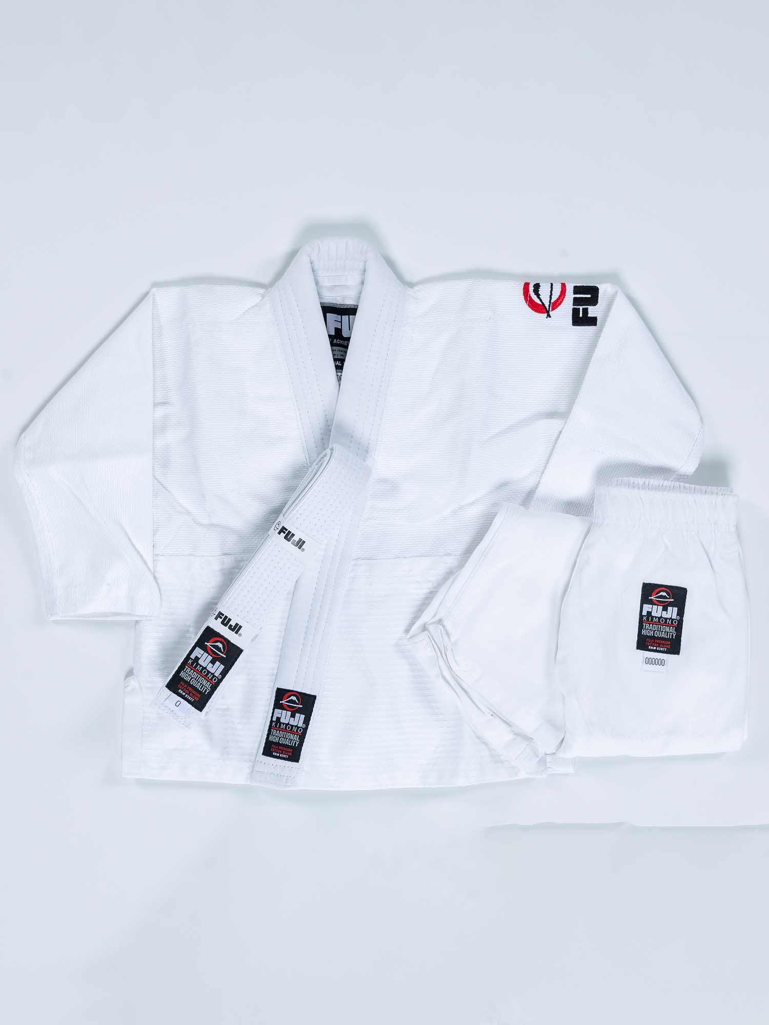 The FUJI Baby Judo Gi in white is shown on a light background, featuring a jacket, pants, and belt with black and red patches plus embroidered logos on the chest, belt, and pant leg.