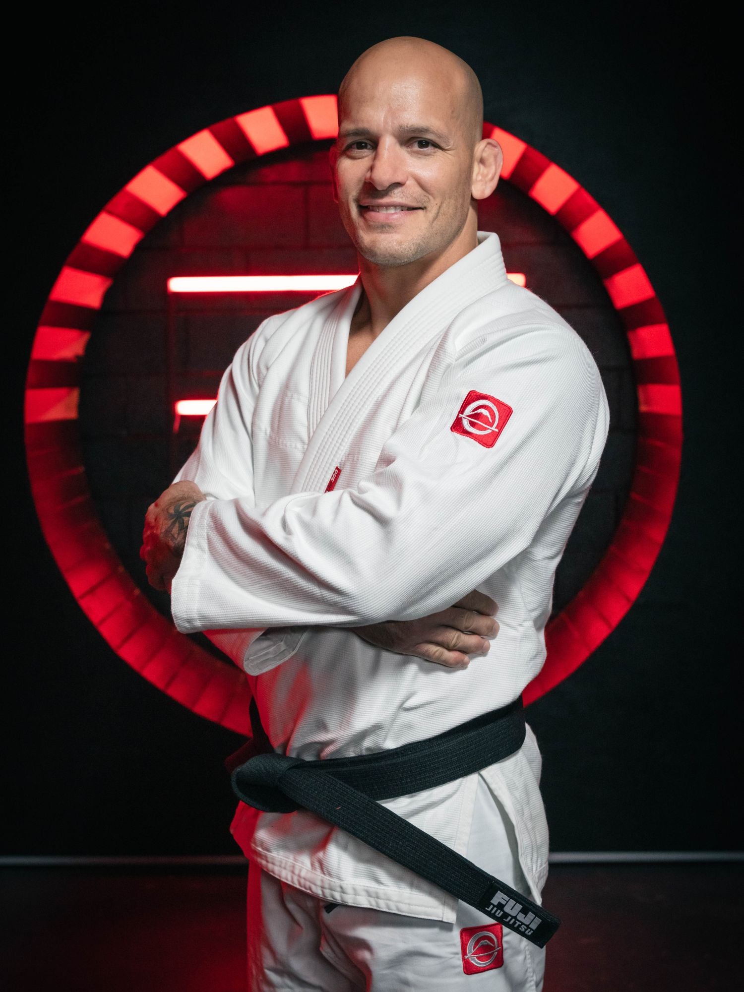 A man wearing the Ultra Lite 300 BJJ Gi White and a black belt stands with arms crossed, smiling in front of a dark background illuminated by a red circular neon light.
