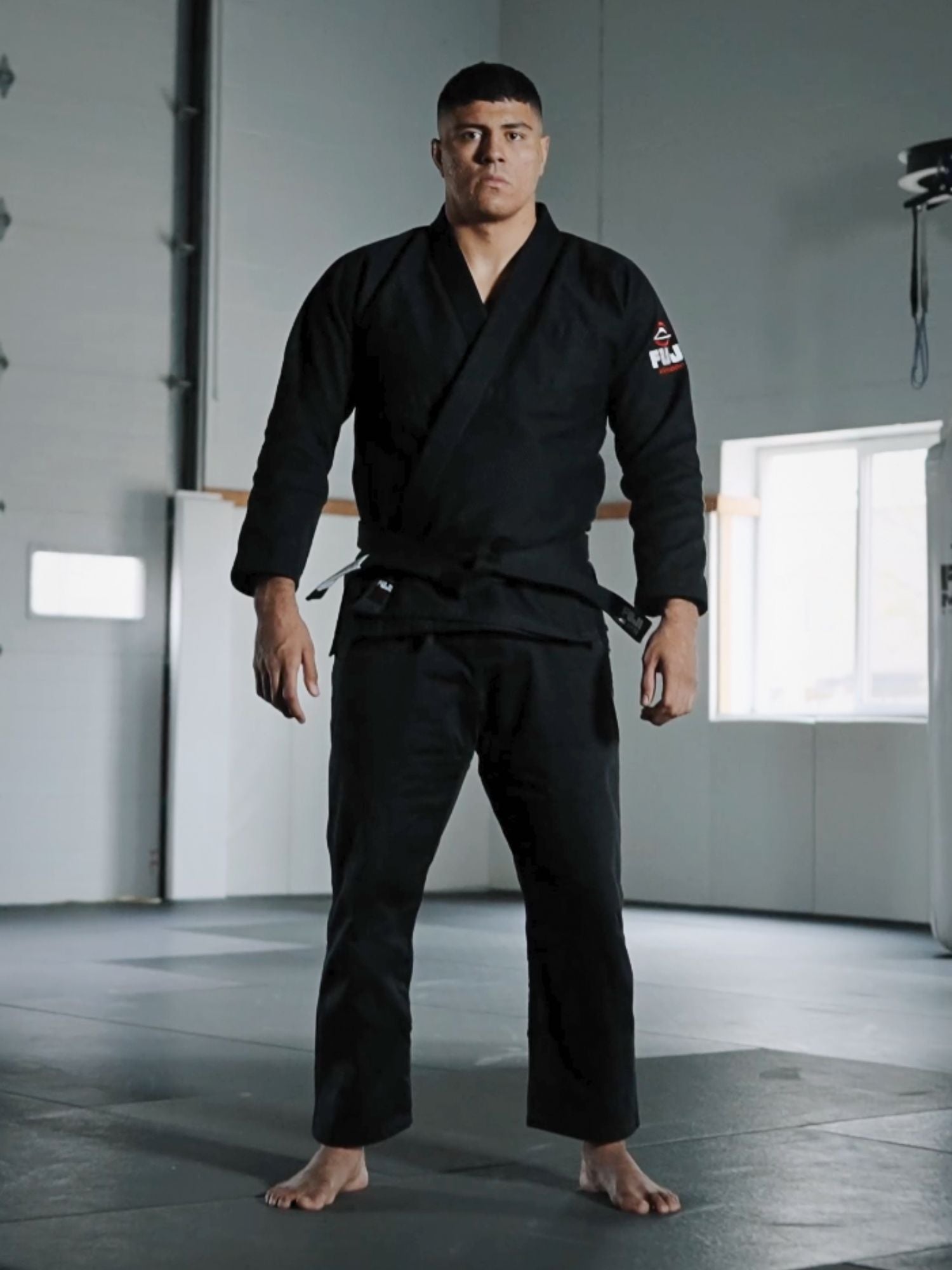 A man stands barefoot on a mat in a well-lit martial arts gym, wearing the All Around BJJ Gi Black and looking directly at the camera.