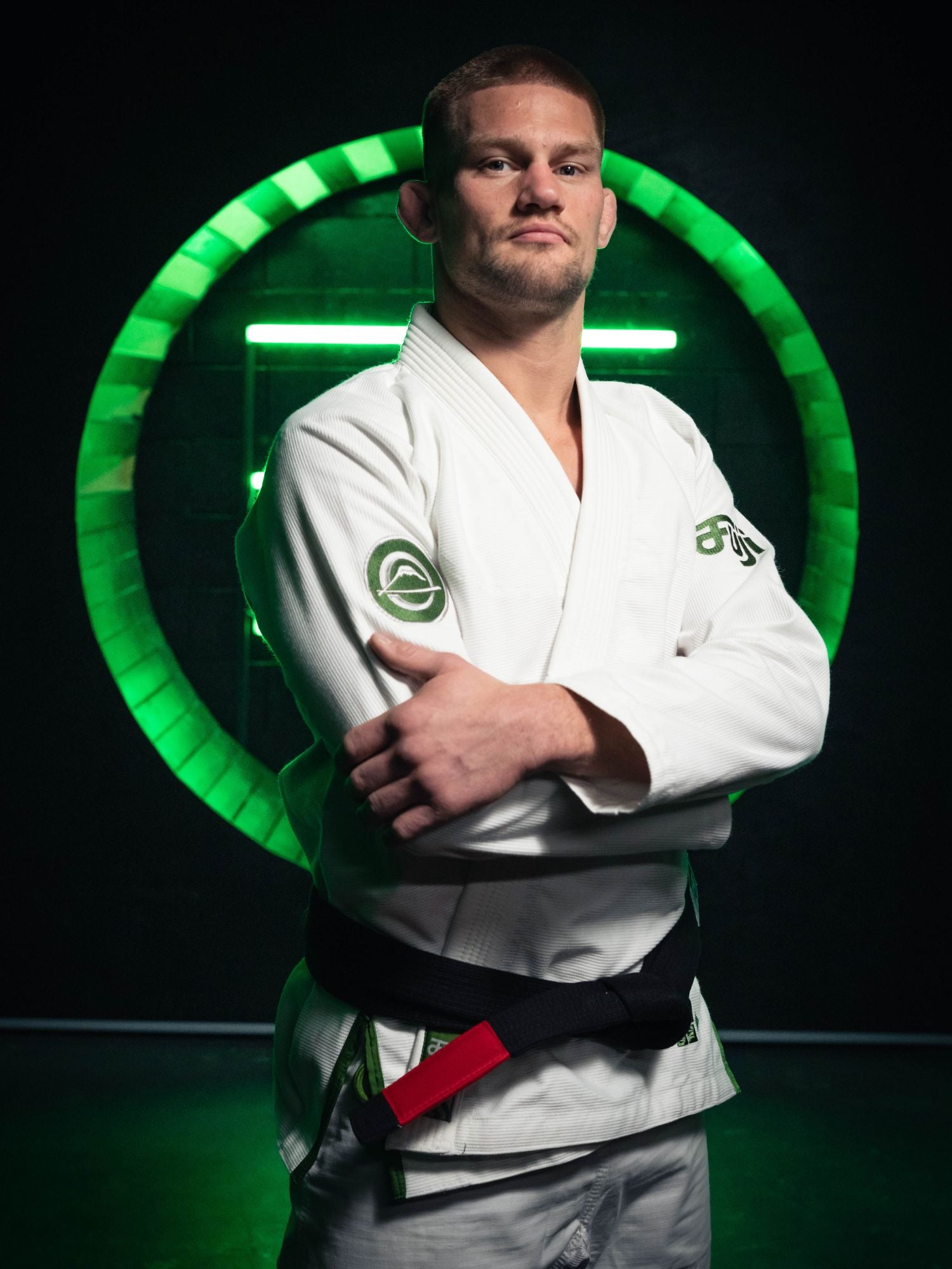 A man wearing the Flow-Tech BJJ Gi White/Green and a black belt stands confidently with arms crossed in front of a glowing green circular neon light against a dark background.