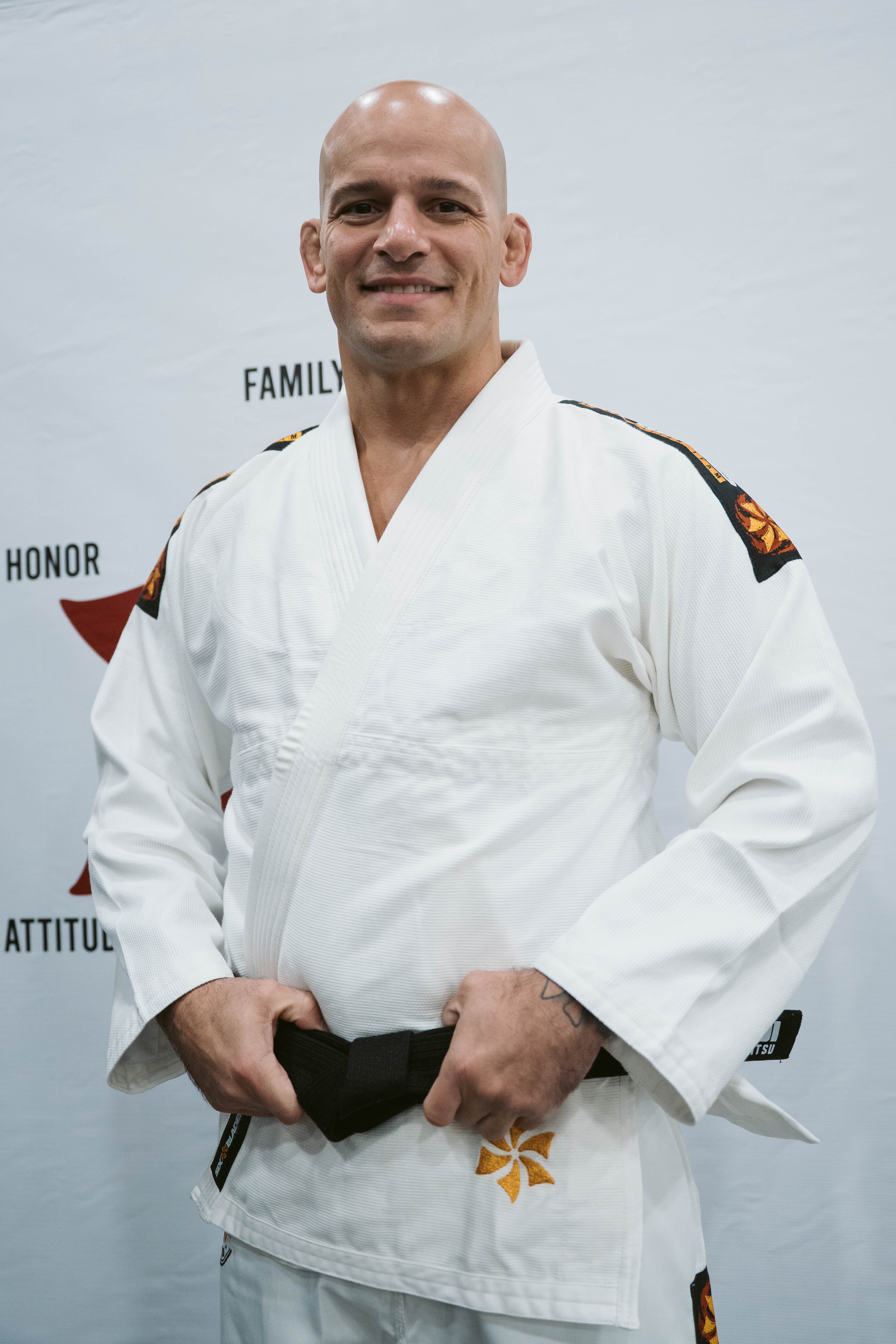 A man wearing the Six Blades Competition Gi with a black belt smiles in front of a light background featuring partially visible motivational words.