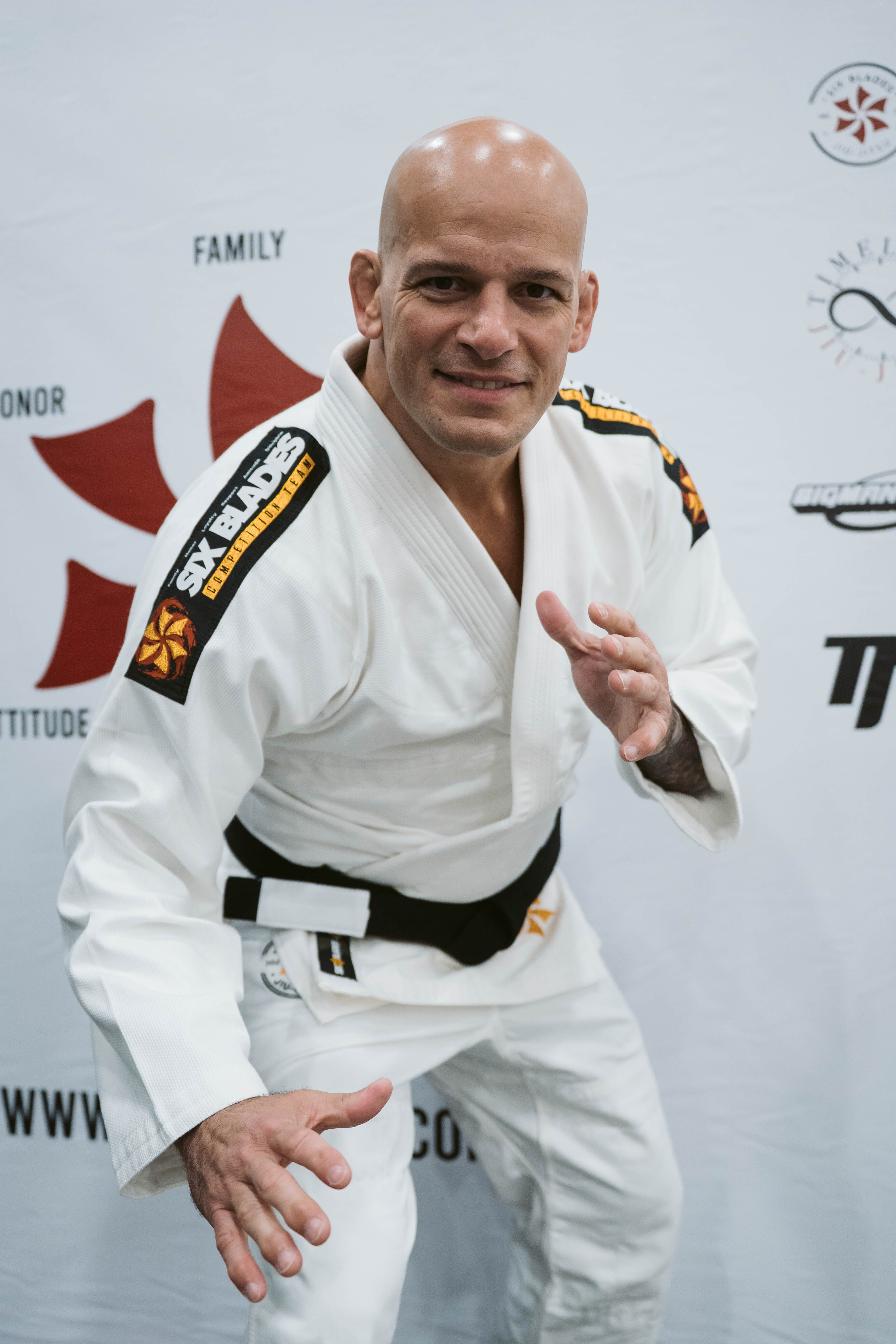 A bald man wearing the Six Blades Competition Gi and black belt poses in a ready stance, smiling, in front of a white backdrop with various logos and text.