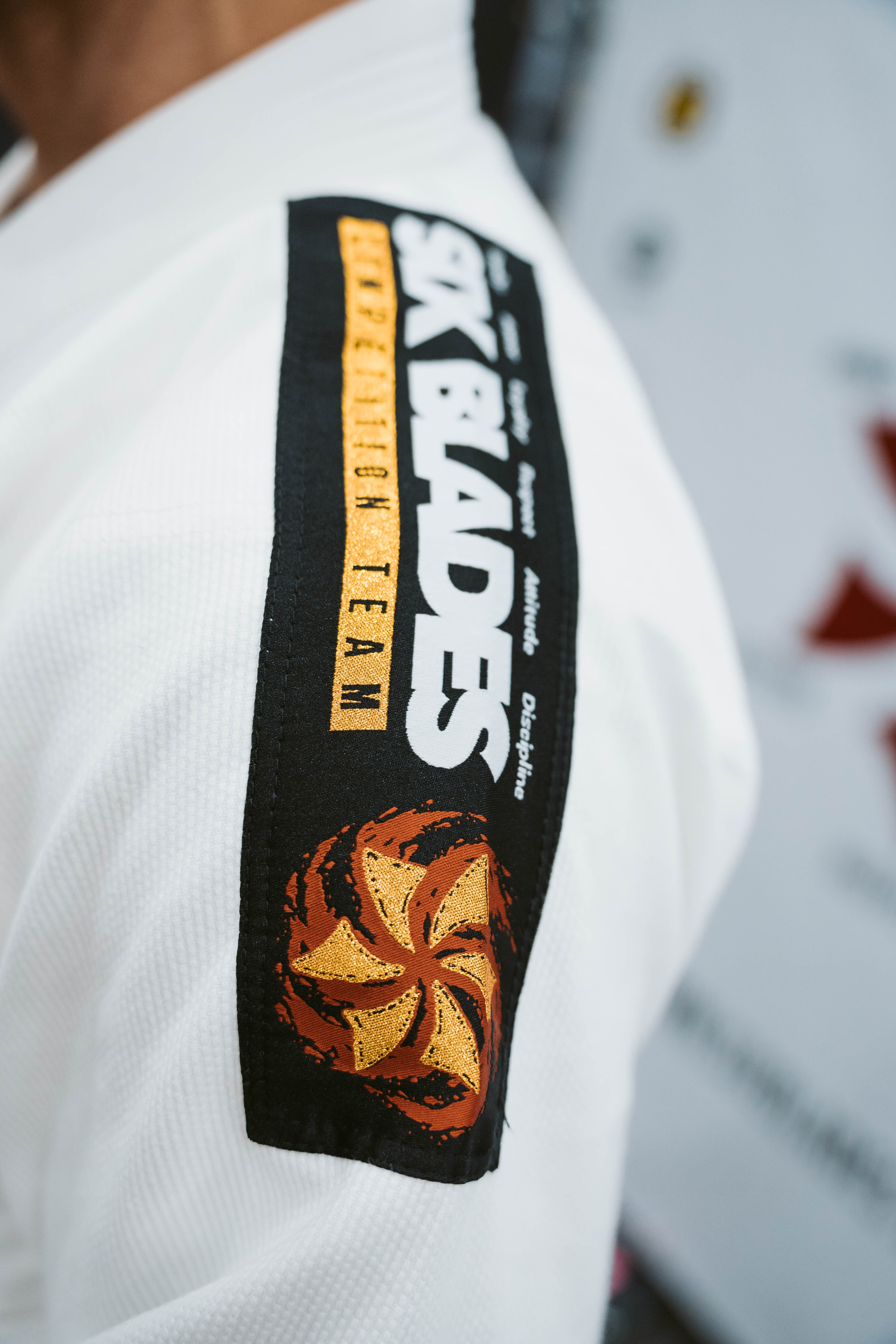 A person wearing the Six Blades Competition Gi, a white martial arts uniform with a black shoulder patch reading "SIX BLADES COMPETITION TEAM" and displaying an orange and yellow emblem.