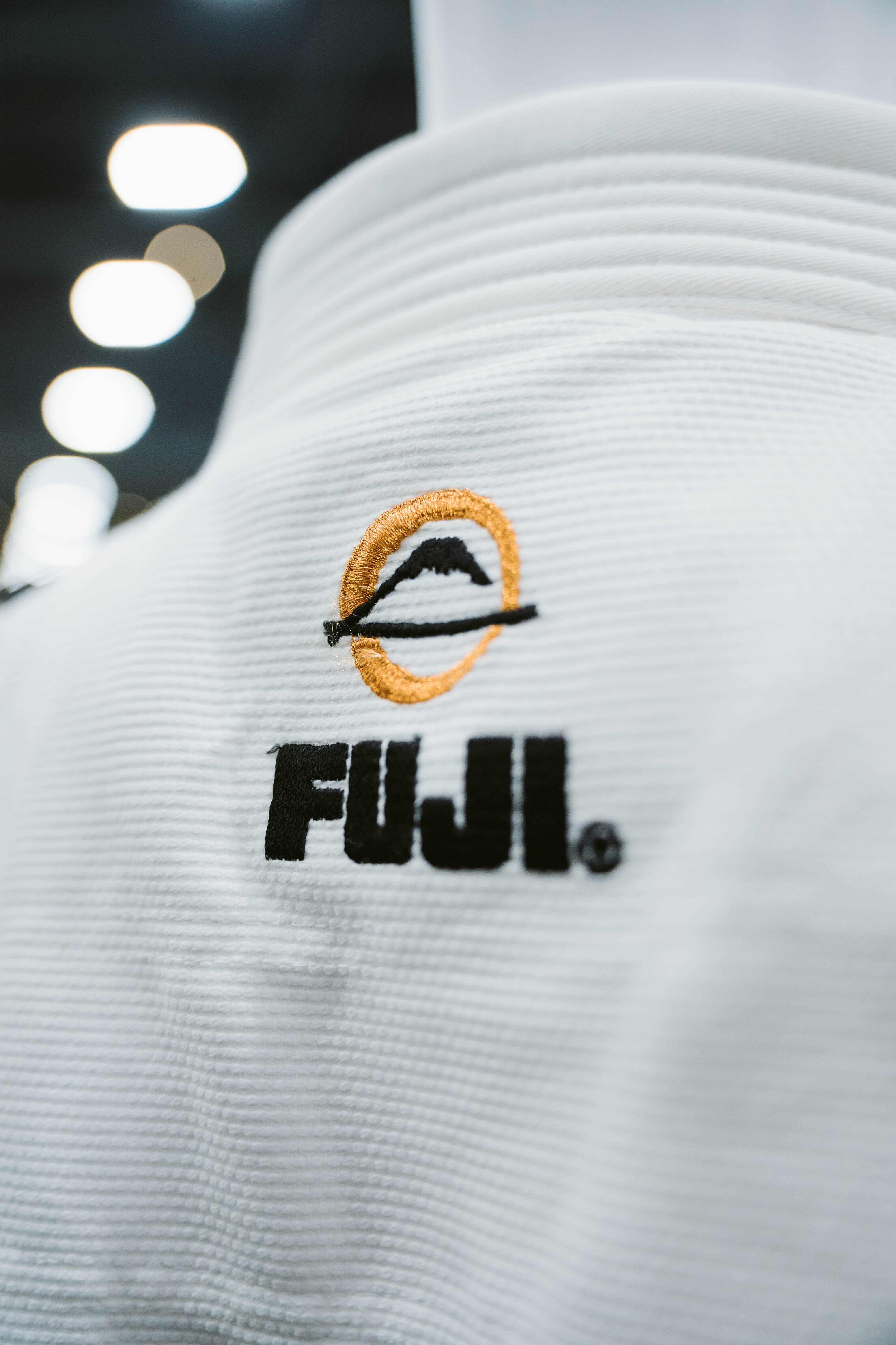 Close-up of the Six Blades Competition Gi, a white martial arts uniform featuring the FUJI logo and an orange mountain symbol embroidered on the fabric. The background is blurred with round lights visible.