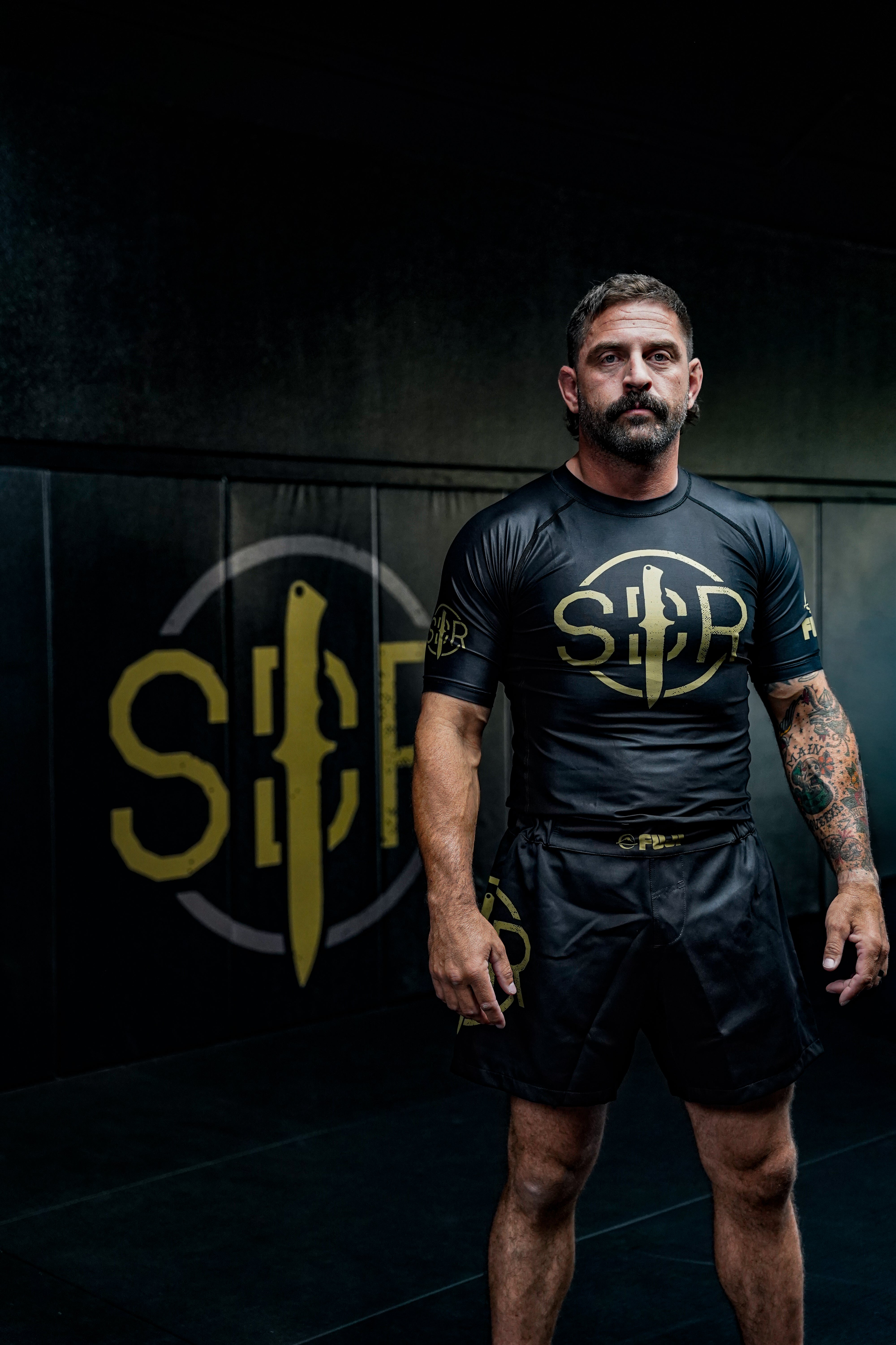 A man with short hair and a beard stands in a gym wearing the Sheepdog Response Essential Short Sleeve Rashguard Black and shorts with yellow logos, with a black wall featuring a large yellow SBF logo in the background.