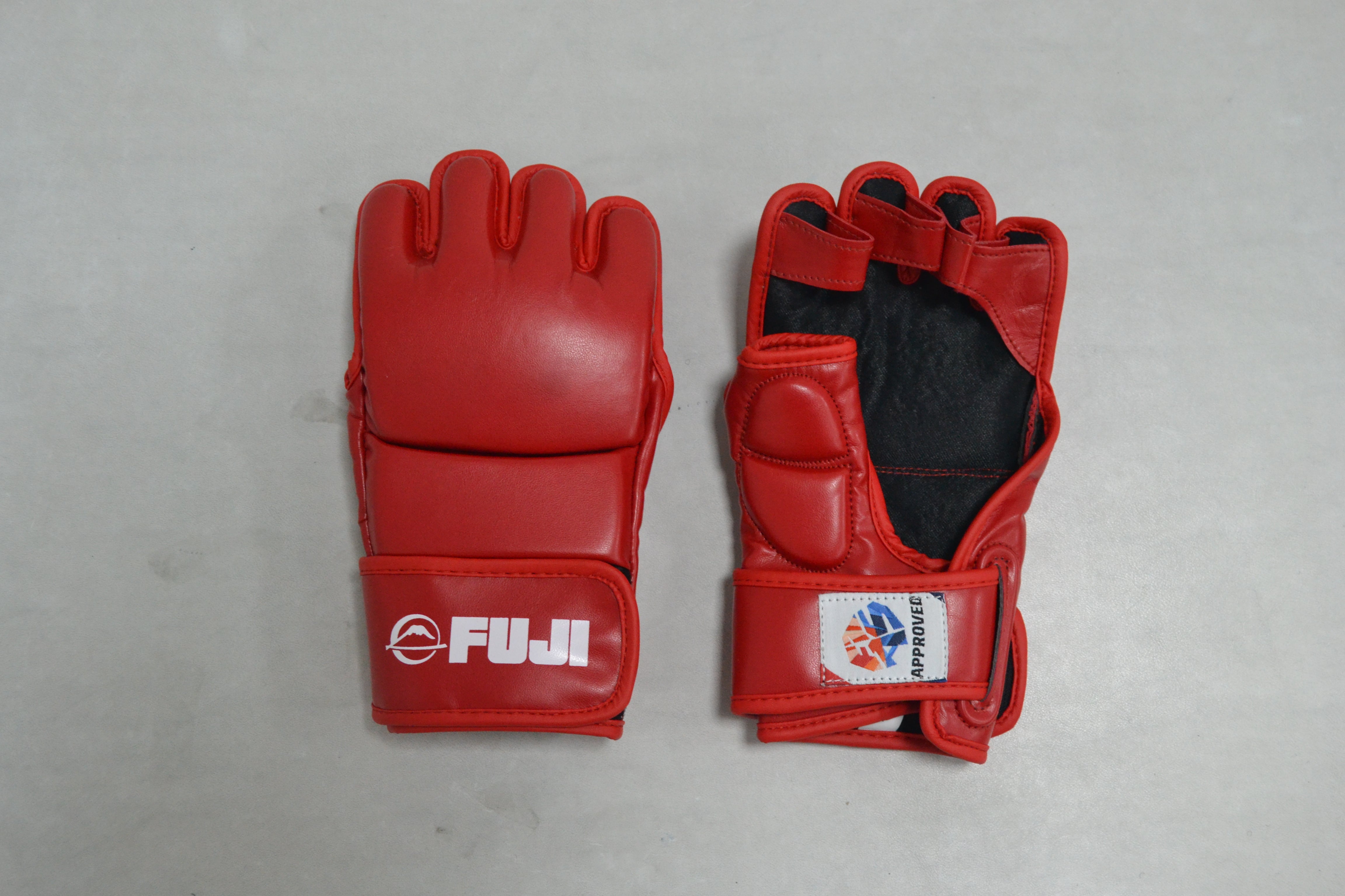 Two FIAS Sambo Combat Gloves in red are displayed on a gray surface. One glove is placed palm down, showcasing the FUJI branding, while the other is palm up, highlighting the open finger design and a small International Federation patch.