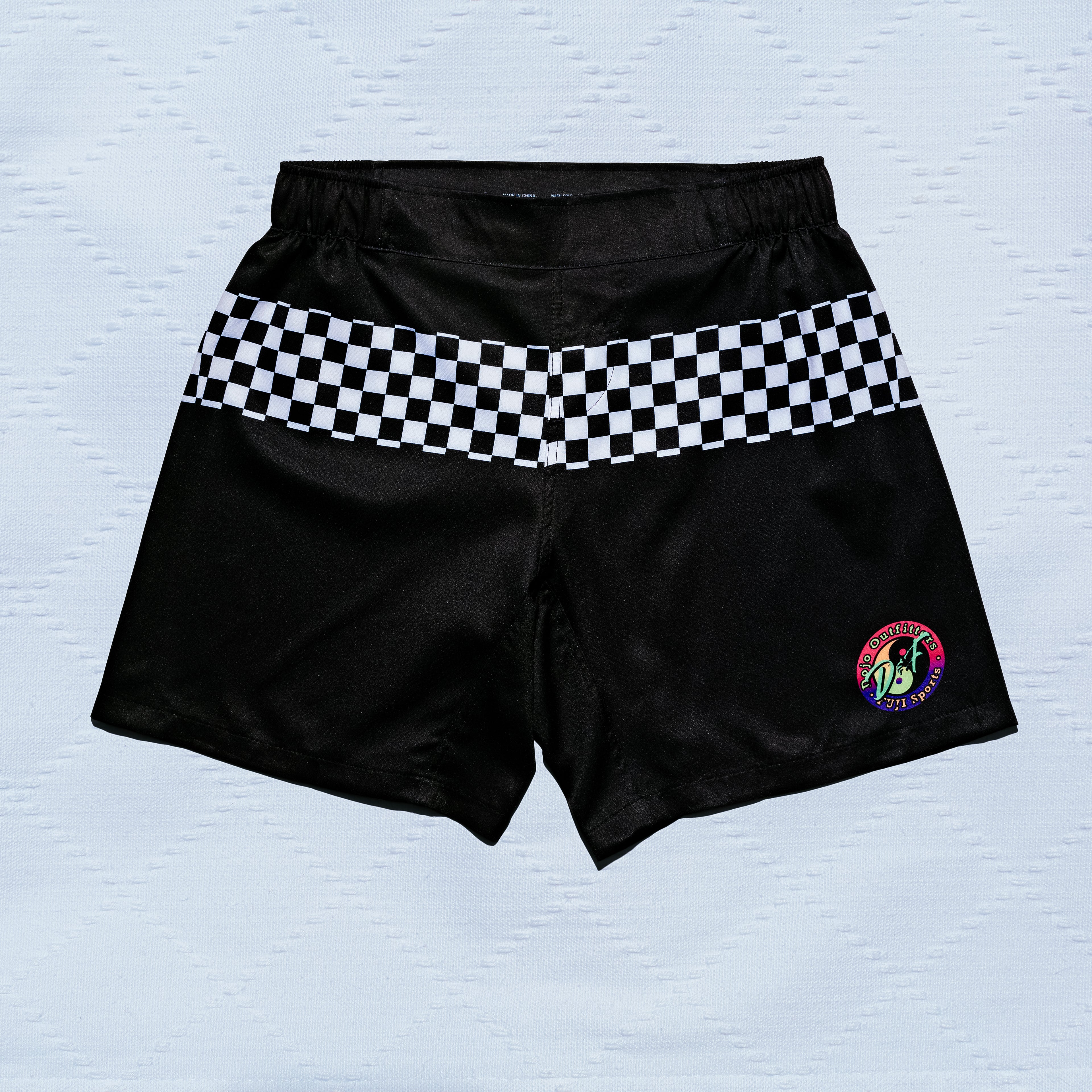 Always Summer DOJO Outfitters Lightweight Shorts in black feature a black and white checkerboard stripe across the front and a vibrant circular "75" patch on the left leg. Displayed on a light, textured surface.