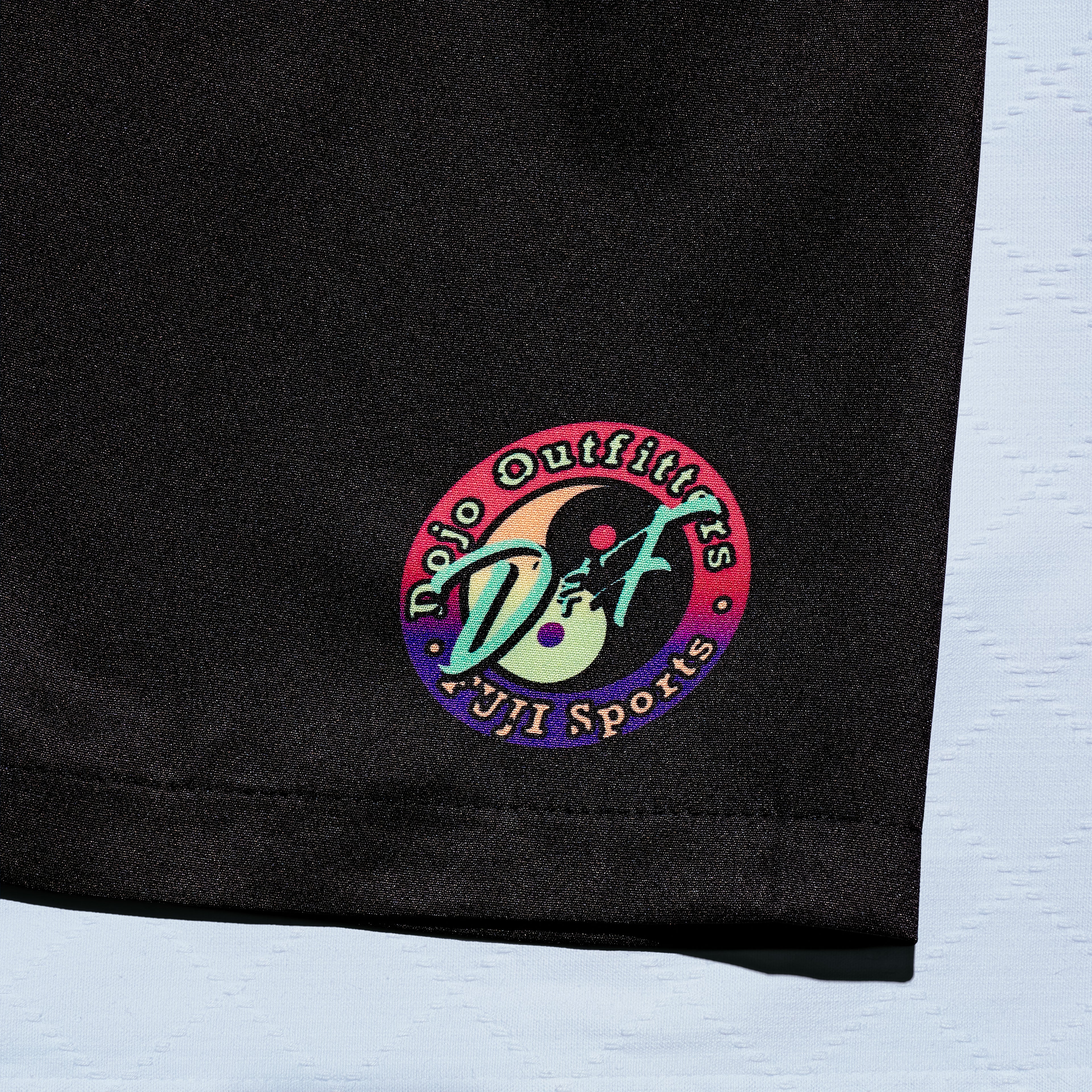 A close-up of the Always Summer DOJO Outfitters Lightweight Shorts shows a black fabric with a vibrant circular “Dojo Outfitters FUJI Sports” patch featuring a martial artist. The background is light blue.