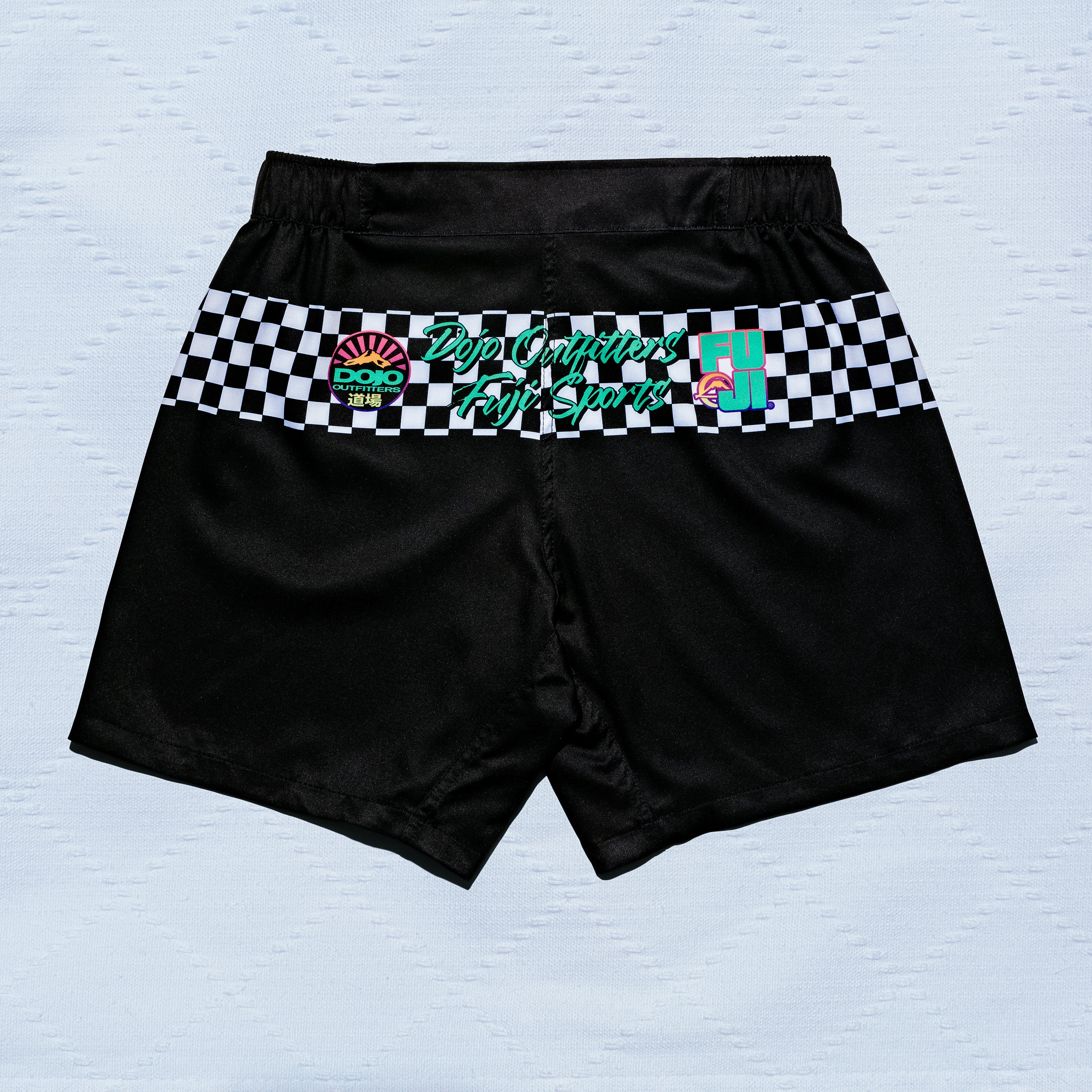 Always Summer DOJO Outfitters Lightweight Shorts in black feature a white and black checkered waistband with colorful logos and "Top Contender Fight Shorts" in green across the back, displayed on a light textured surface.
