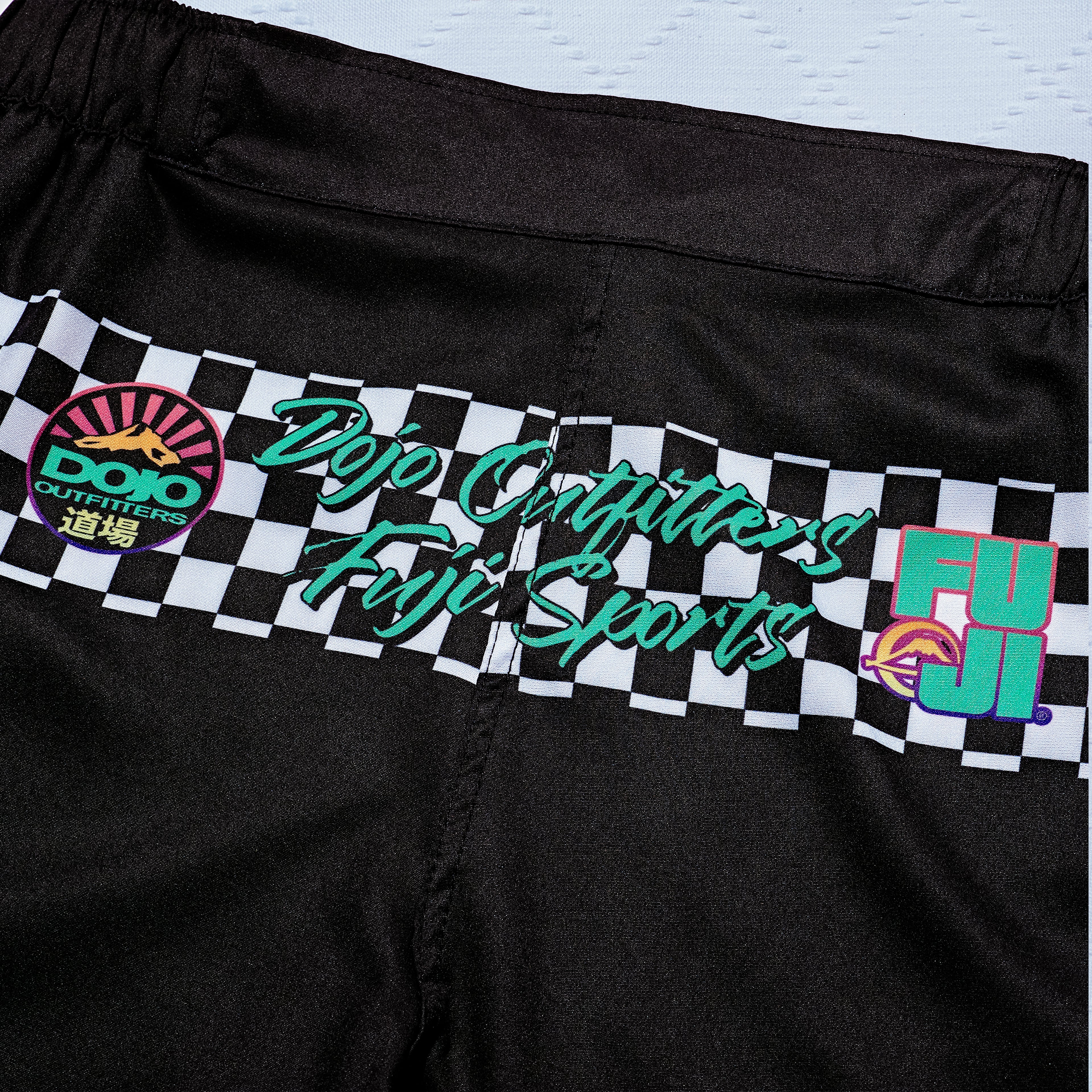 The Always Summer DOJO Outfitters Lightweight Shorts are black athletic shorts with a black-and-white checkerboard stripe and colorful logos: Dojo Outfitters, Fuji Sports, and a purple FUJI with a mountain graphic.