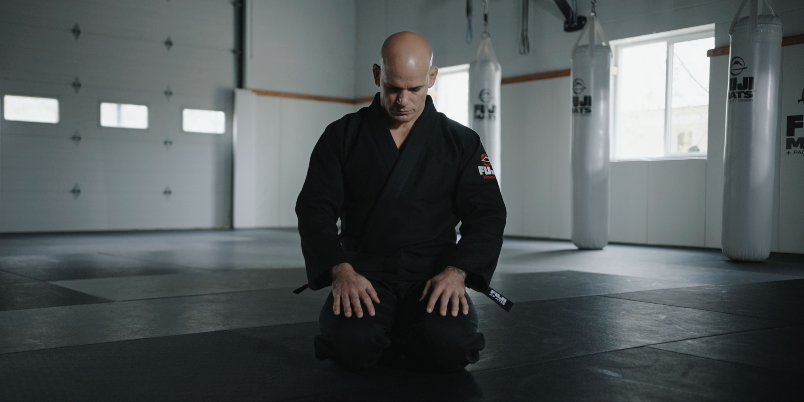A man in a black martial arts gi kneels on a mat in a quiet, sunlit dojo, head bowed and hands resting on his thighs, surrounded by white punching bags and large windows.
