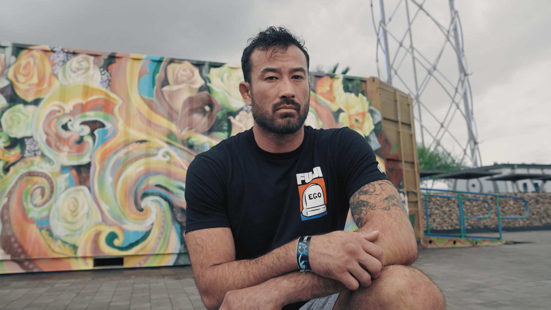 A man with short dark hair and a beard sits in front of a colorful mural, wearing a black shirt and displaying tattoos on his arm. The background includes a container and a metal structure.