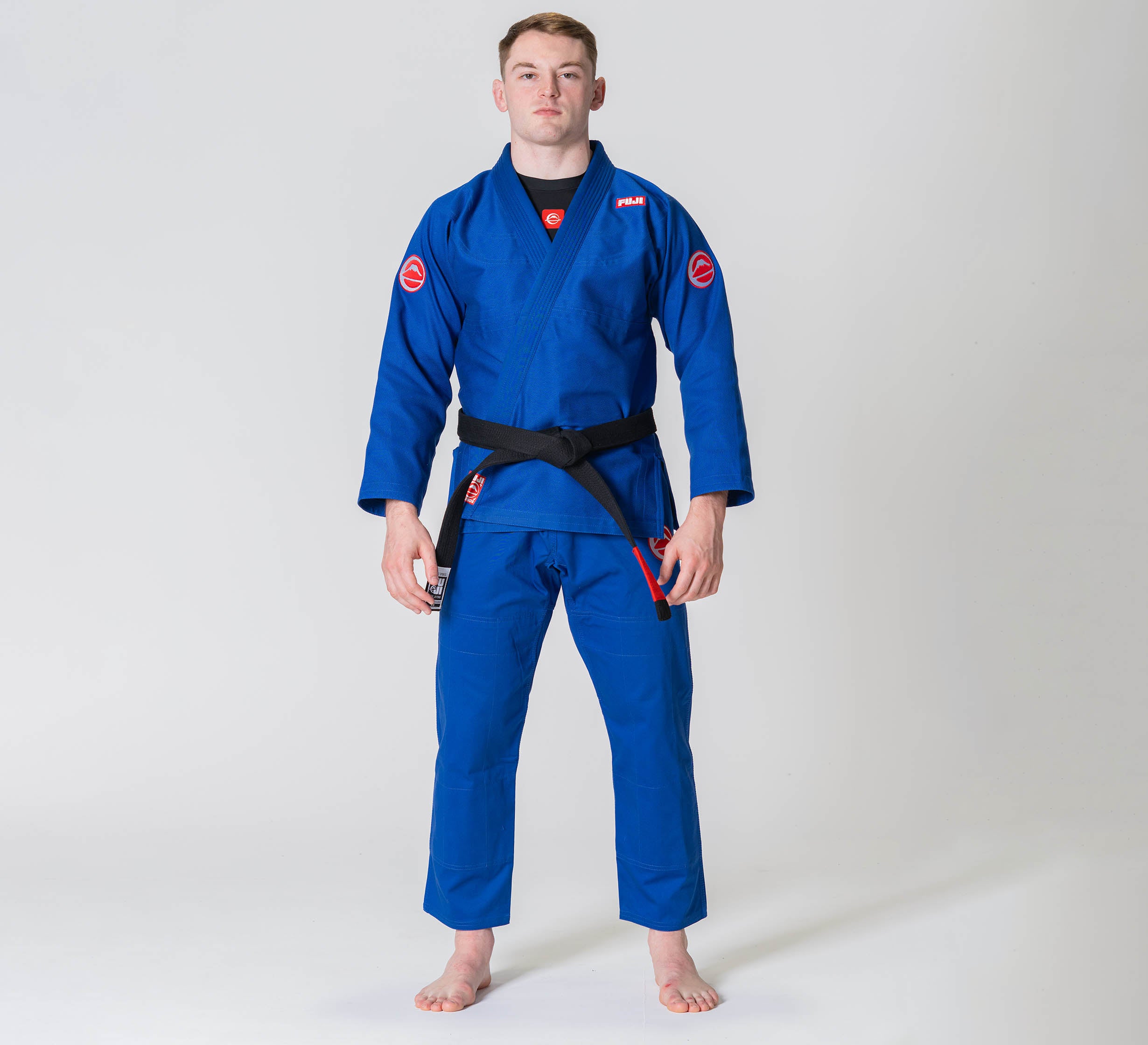 A man stands barefoot on a white background, wearing the Competition BJJ Gi Blue and a black belt. He faces forward with a neutral expression, arms relaxed at his sides.