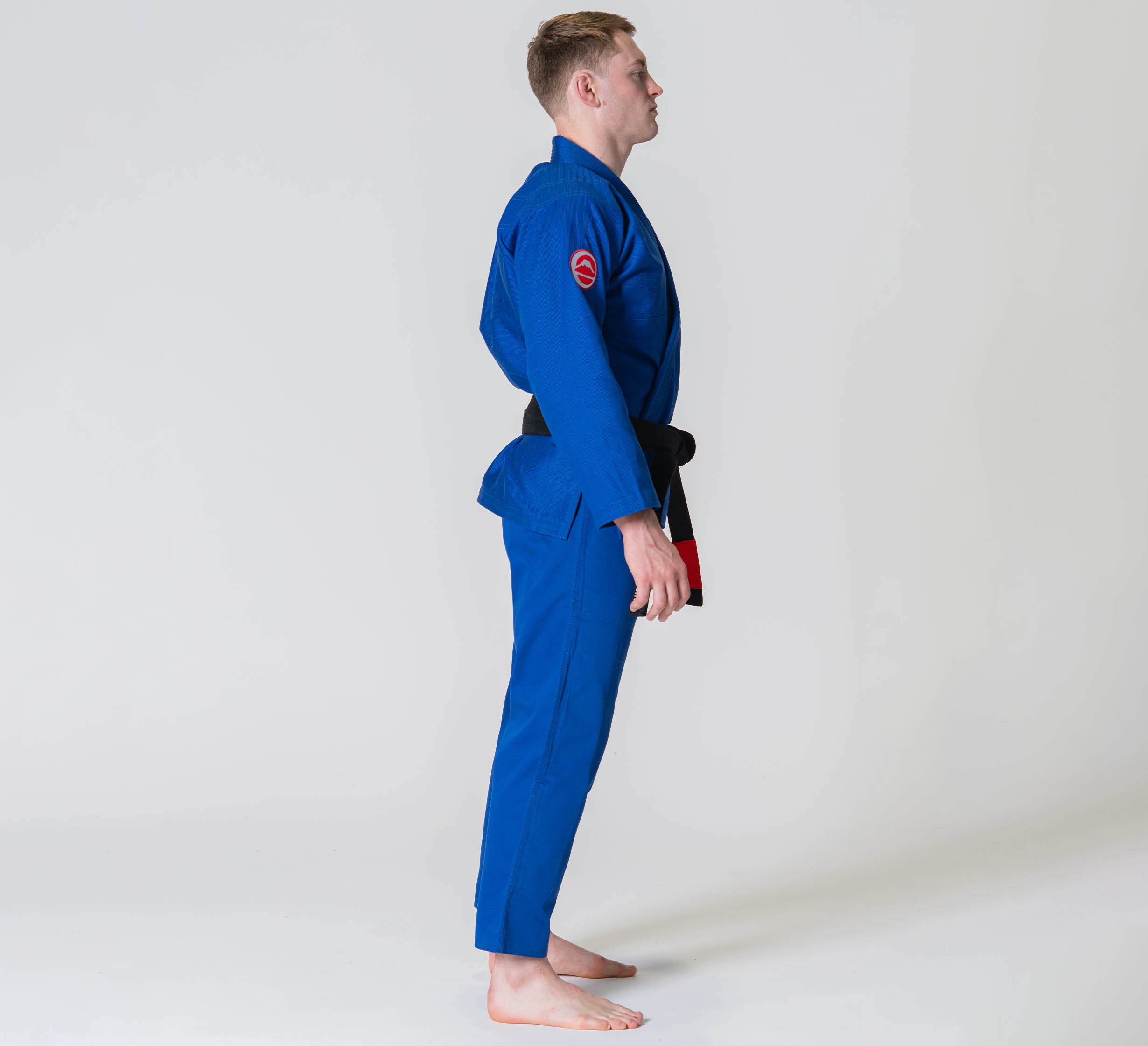 A person wearing the Competition BJJ Gi Blue and a black belt stands barefoot in a side profile pose against a plain light background.