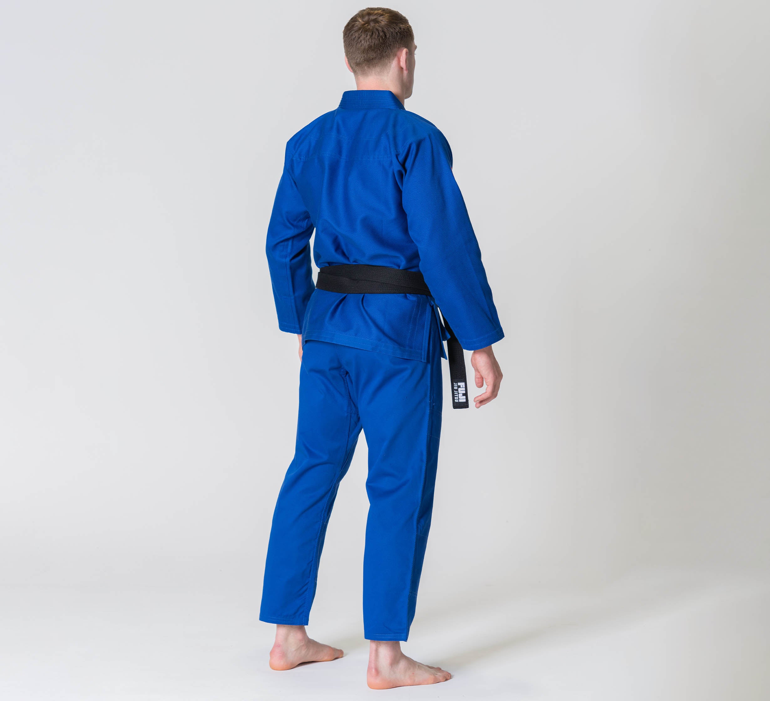 A person wearing the Competition BJJ Gi Blue and a black belt stands barefoot, facing away, against a plain light background.