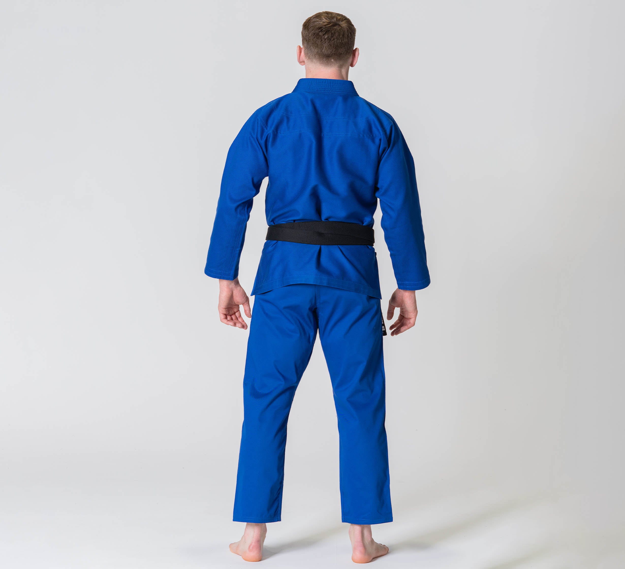A person stands barefoot on a white background, seen from behind, wearing the Competition BJJ Gi Blue and a black belt.