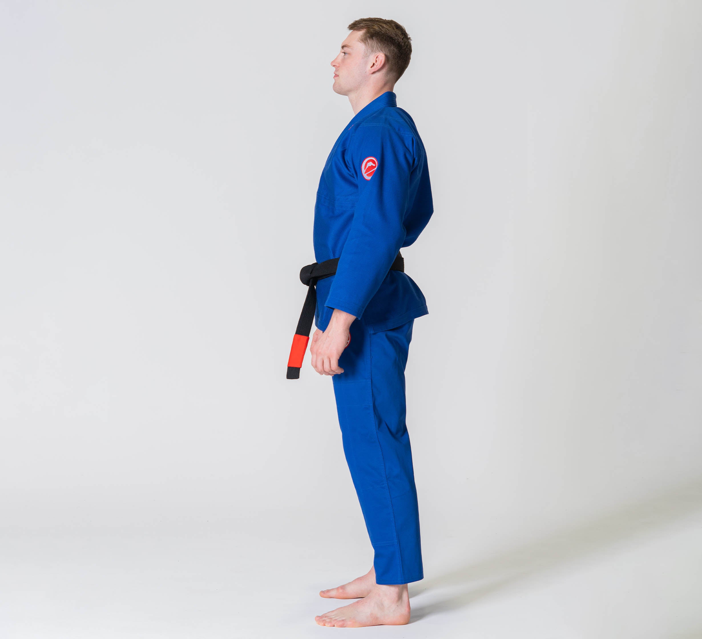 A person stands barefoot in profile against a plain white background, wearing the Competition BJJ Gi Blue with a black belt.
