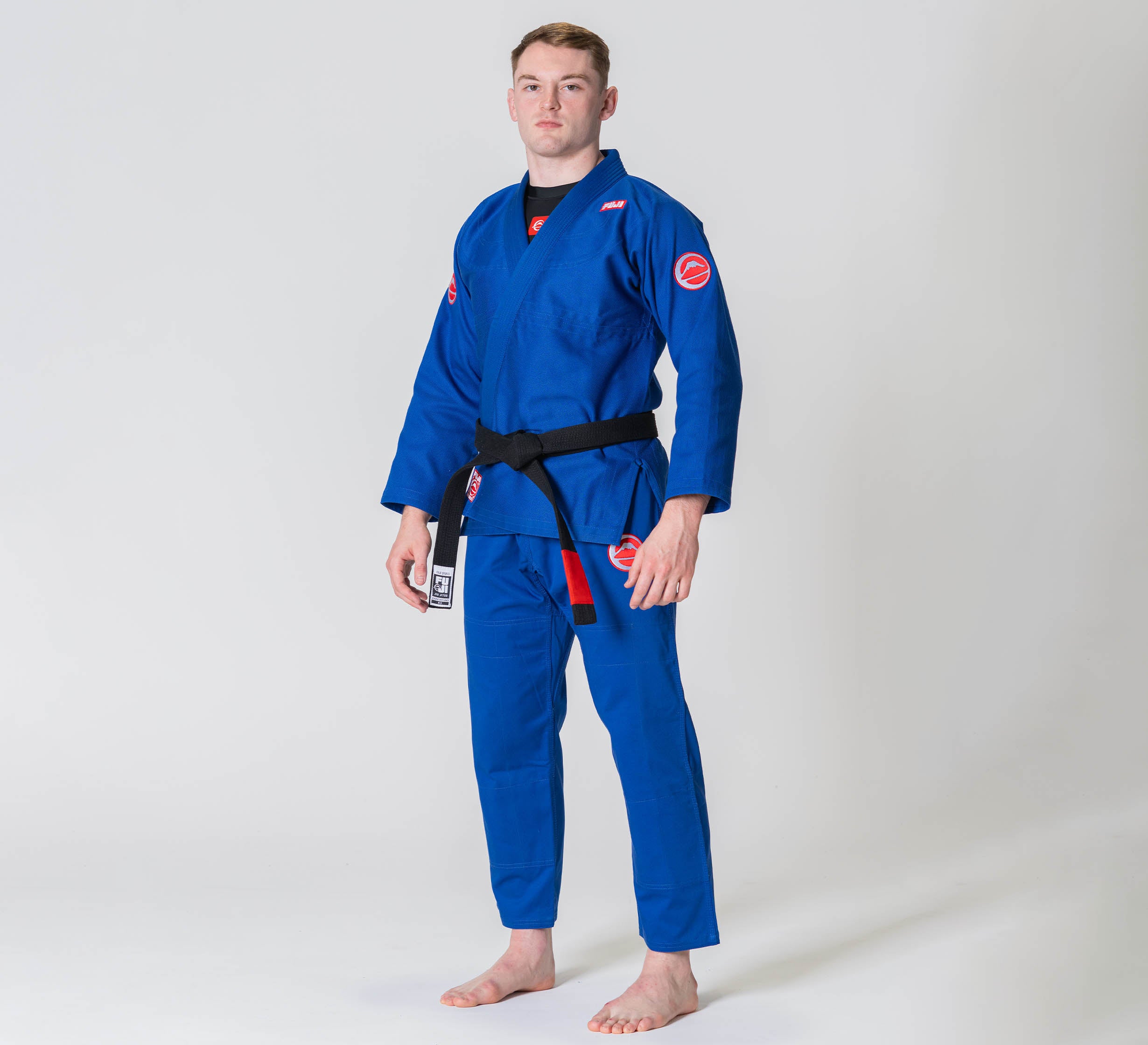 A person stands barefoot on a white background, wearing the Competition BJJ Gi Blue with a black belt. The gi features red patches on the shoulders and sleeves. The individual faces forward with a neutral expression.