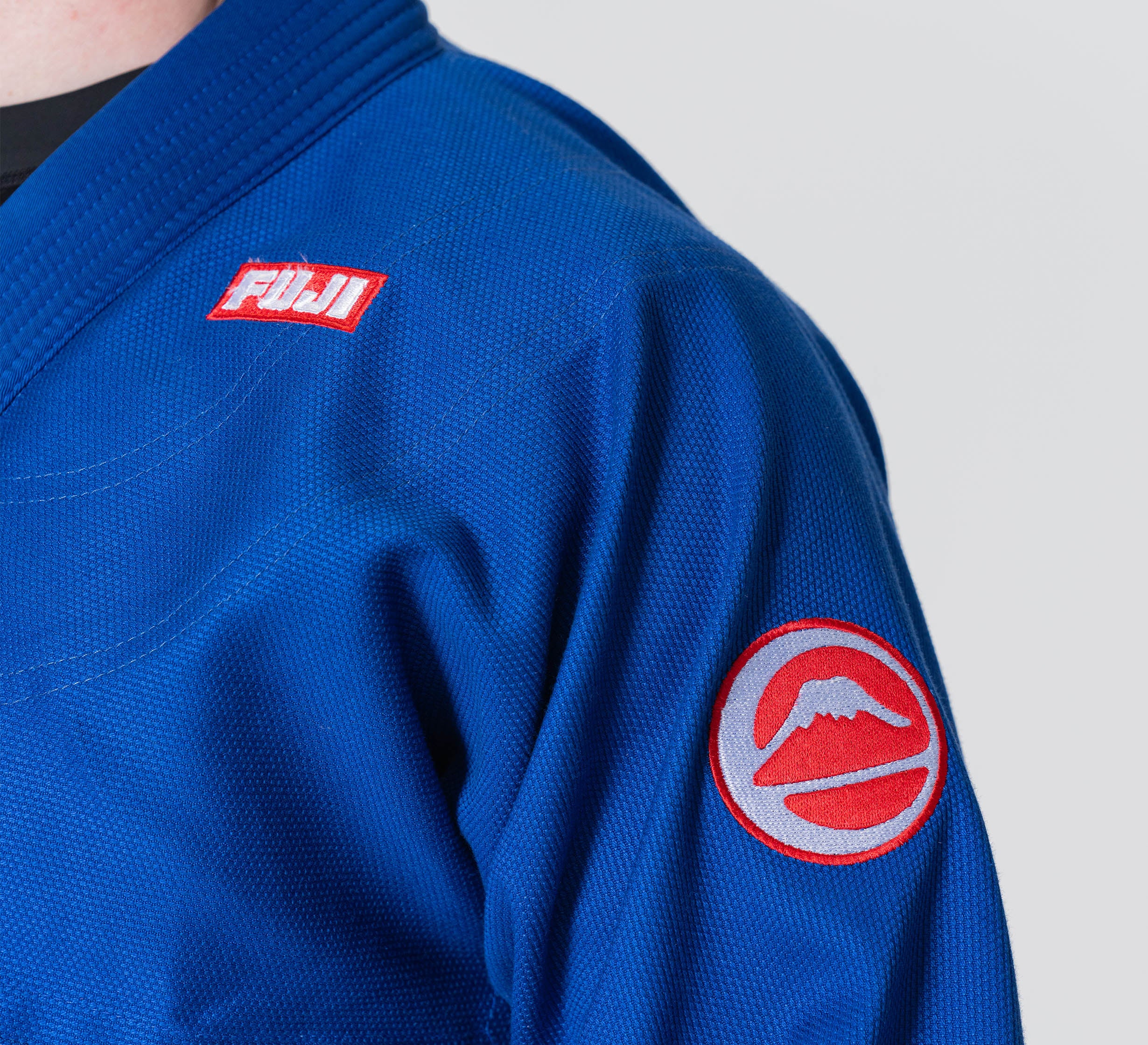 A person wearing the Competition BJJ Gi Blue, featuring a Fuji logo patch on the chest and a red circular mountain patch on the upper sleeve.