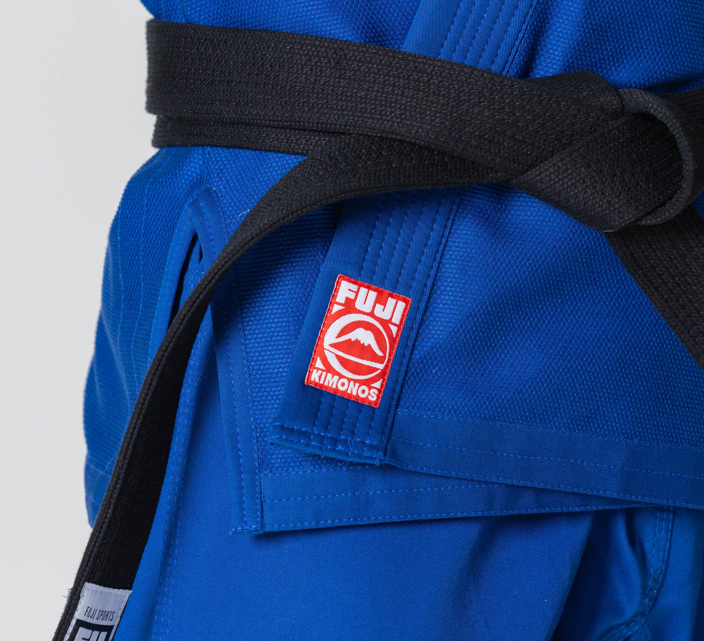 Close-up of a person wearing the Competition BJJ Gi Blue with a black belt. The gi jacket features a red and white Fuji Kimonos patch.