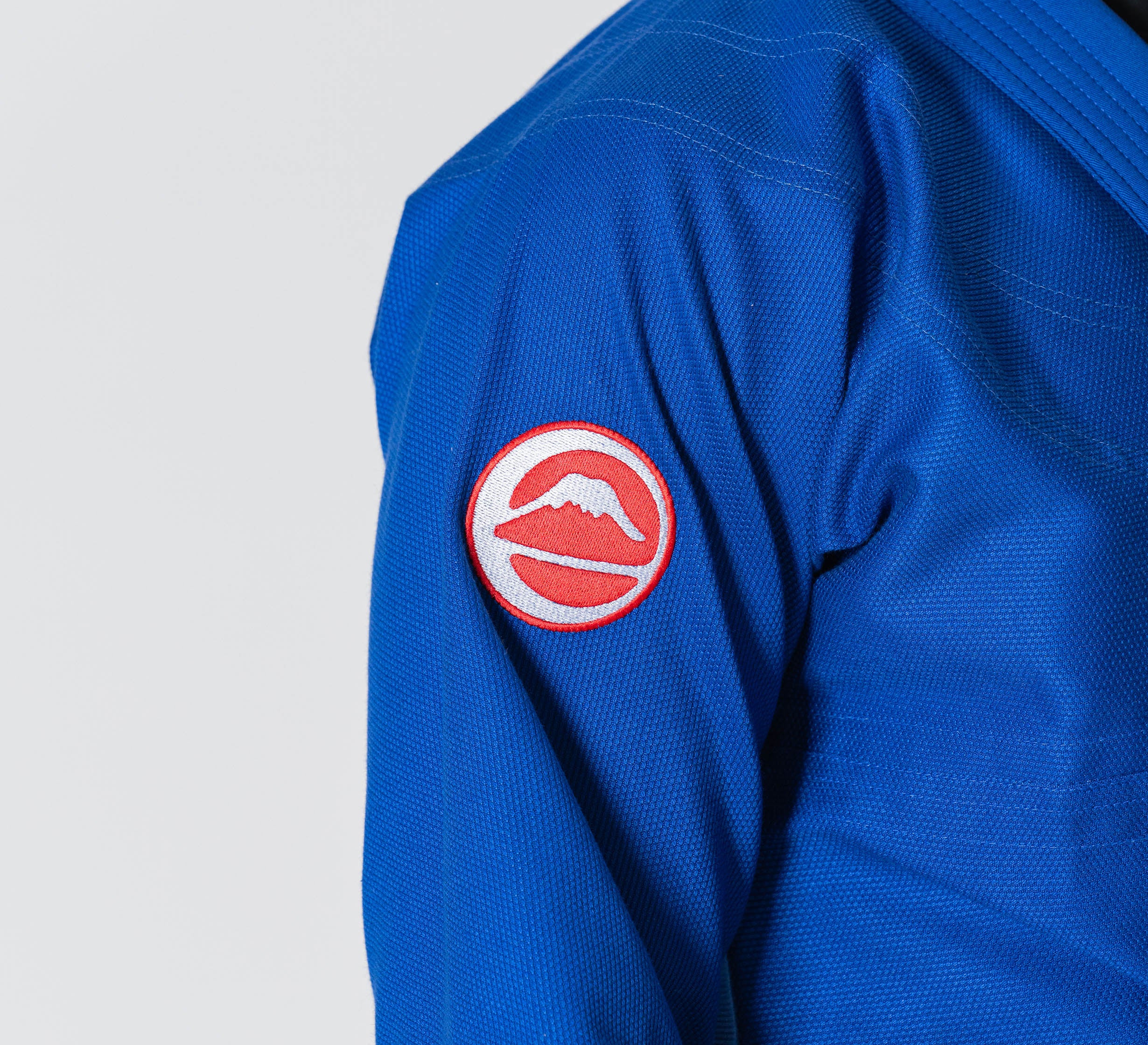 Person wearing the Competition BJJ Gi Blue, featuring a round patch with a white mountain over a red background on the sleeve. The image is cropped to show only the upper body and arm.