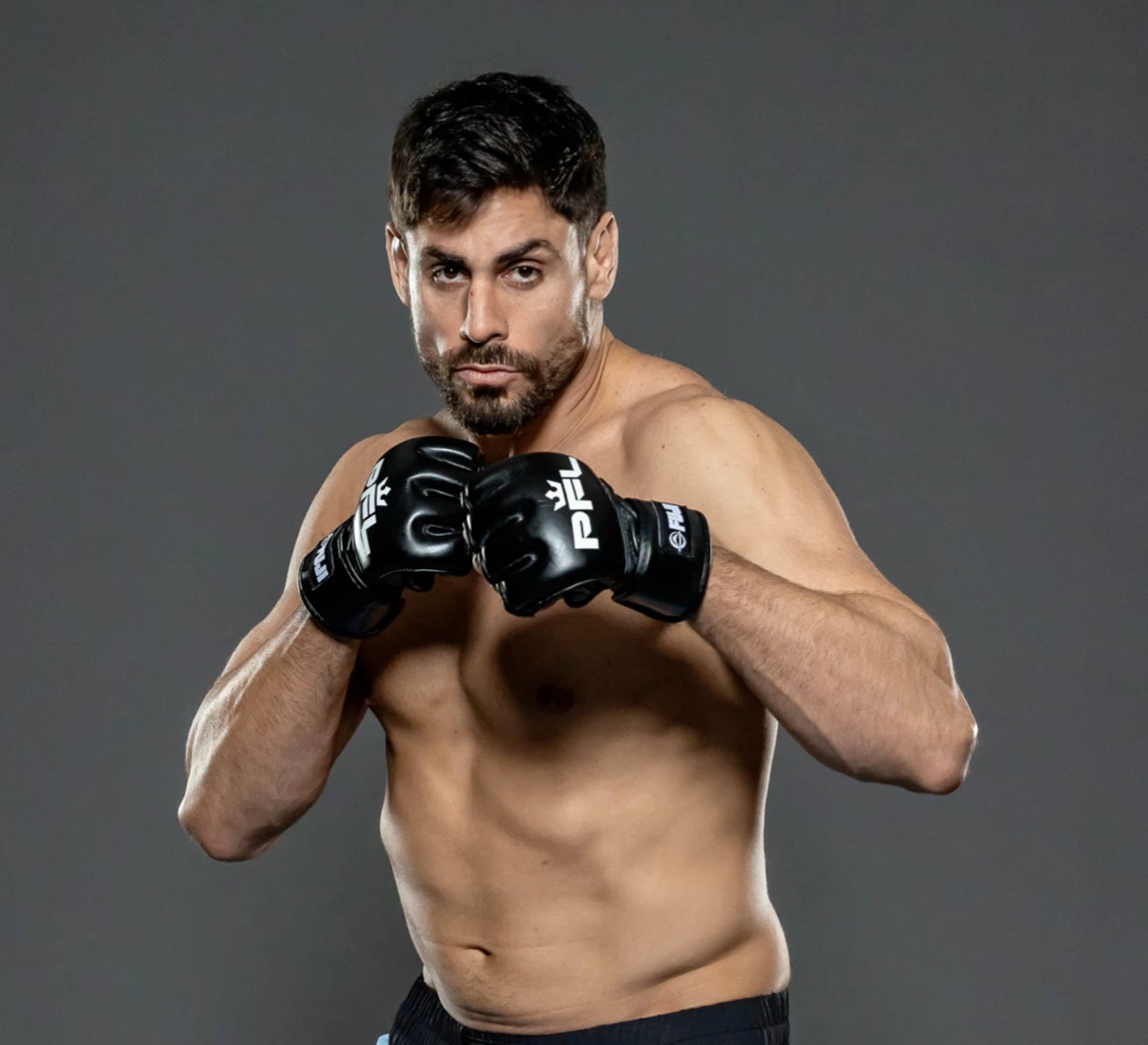 A male mixed martial artist poses shirtless against a gray background, wearing PFL MMA Gloves Black with fists raised in a fighting stance and a focused, determined expression.