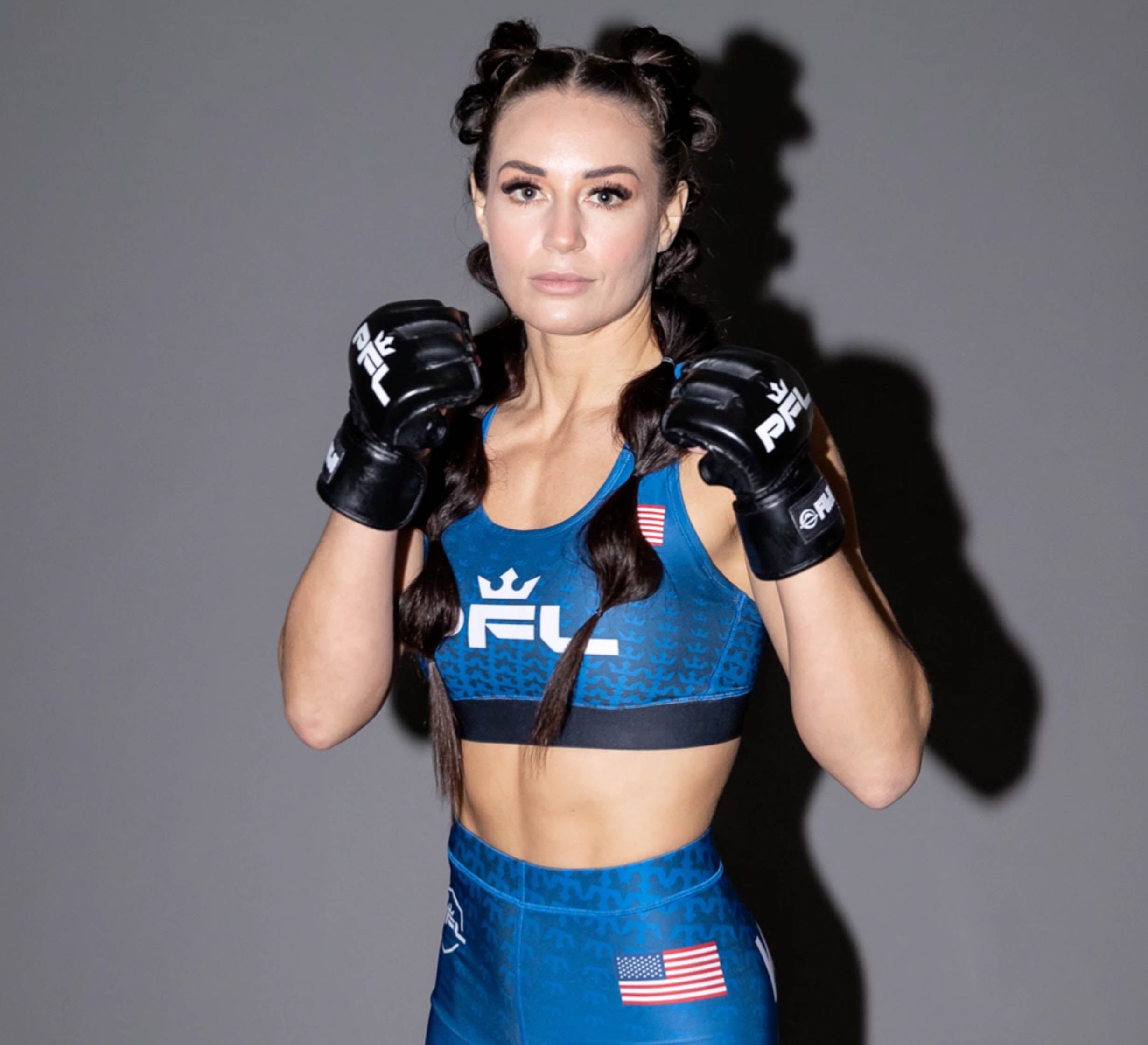A female MMA fighter in blue PFL gear and PFL MMA Gloves Black stands with fists raised in a fighting stance, confidently facing the camera. An American flag patch is shown on her shorts.