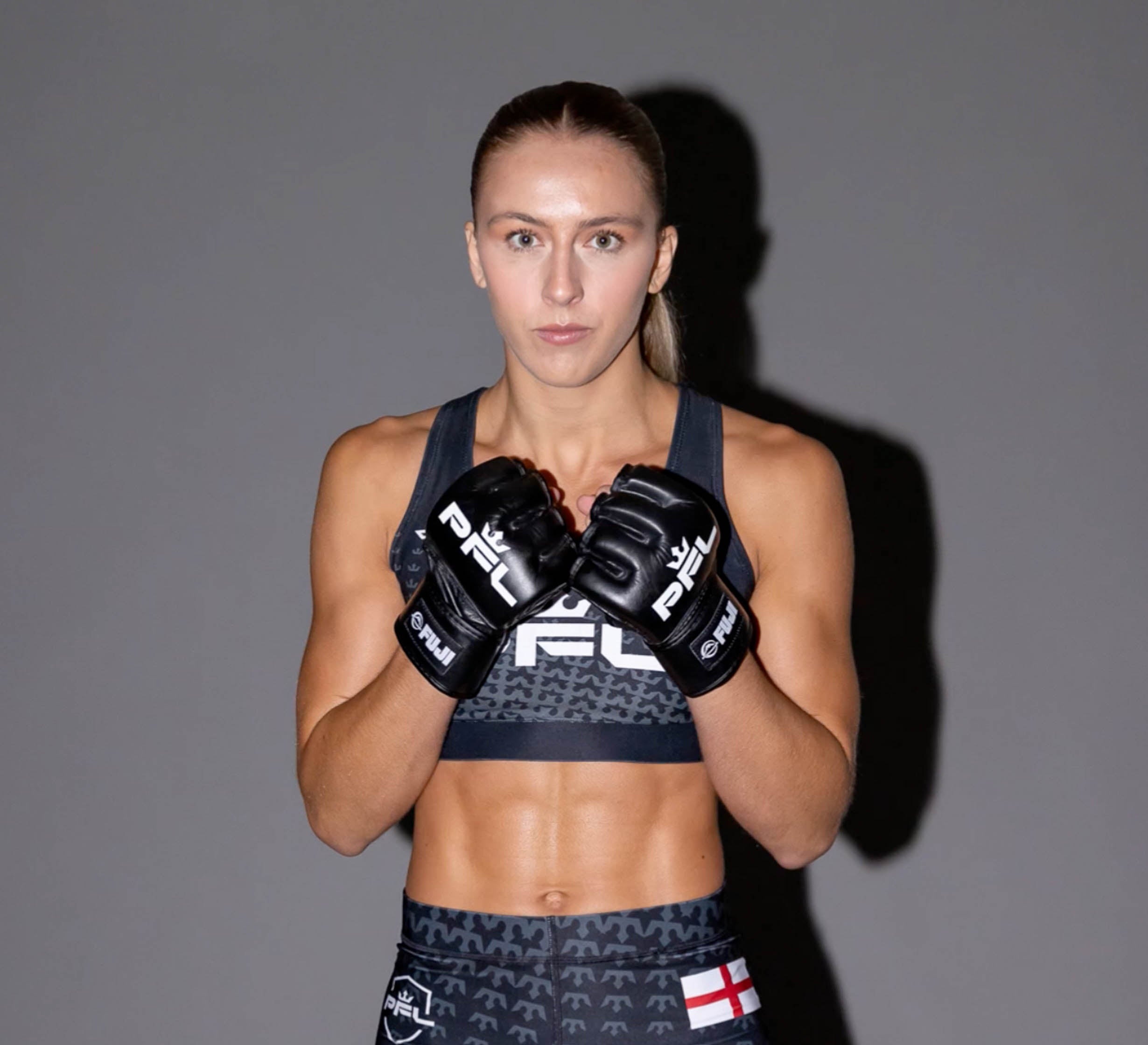 A female mixed martial artist poses in a fighting stance, wearing PFL MMA Gloves Black and a matching sports outfit with the PFL logo and an England flag patch, set against a plain gray background.