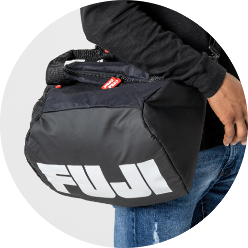 A person wearing blue jeans and a black shirt holds a black FUJI sports bag with a white logo and a visible red zipper pull, against a plain white background.