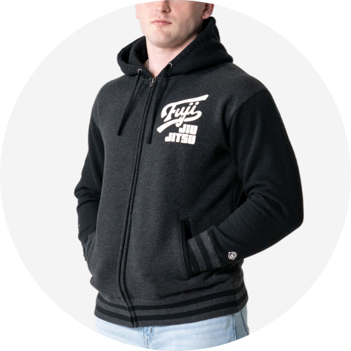 A person wearing a black and dark gray zip-up hoodie with Fuji Jiu Jitsu printed in white on the left chest, standing with hands in pockets against a plain light background.