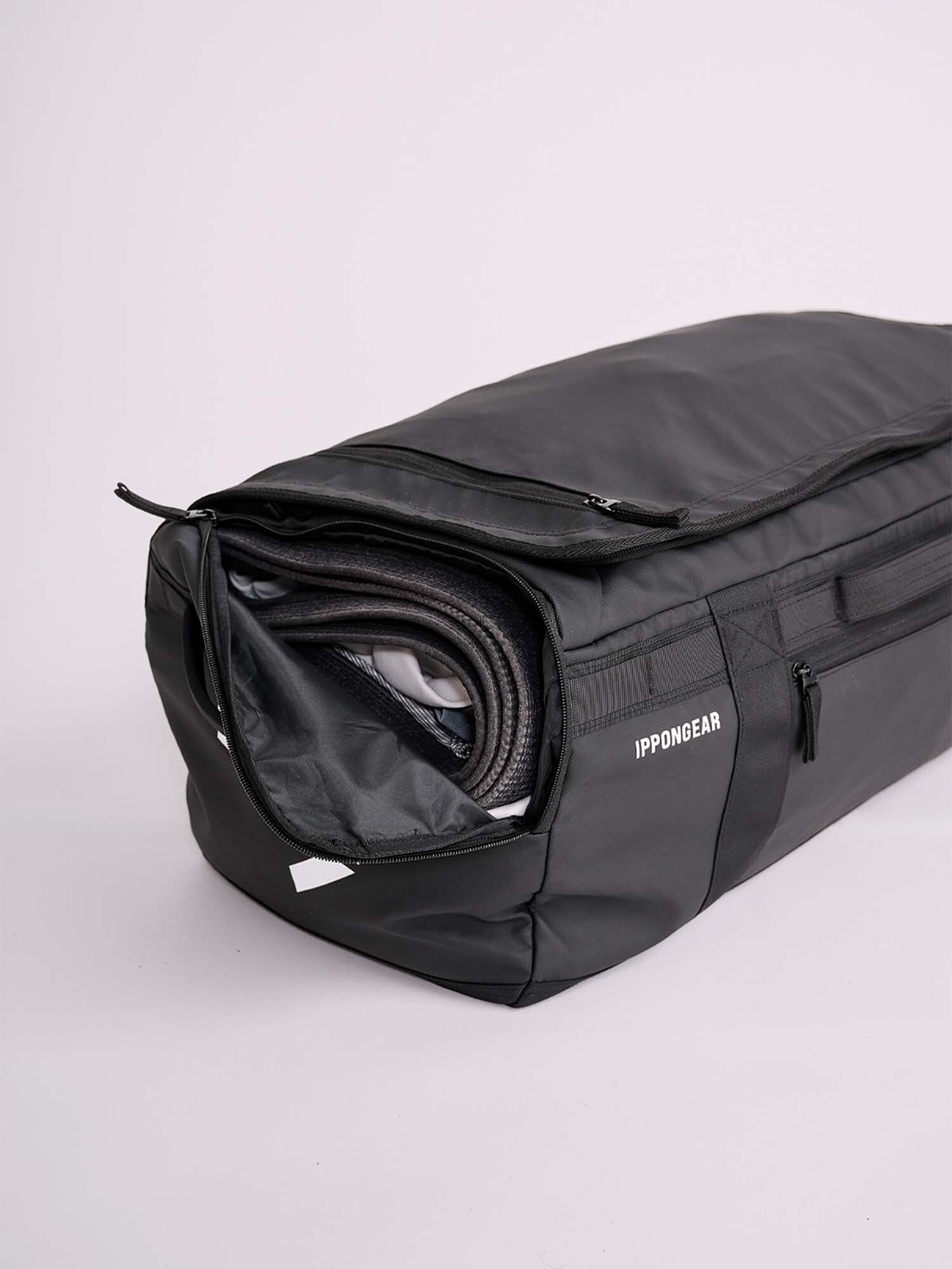 The IPPONGEAR 2 in 1 Sports Bag Black is a spacious duffel with a partially open zipper, showing a rolled-up martial arts belt inside. The IPPONGEAR logo is on the side, and the bag is set against a simple light background.