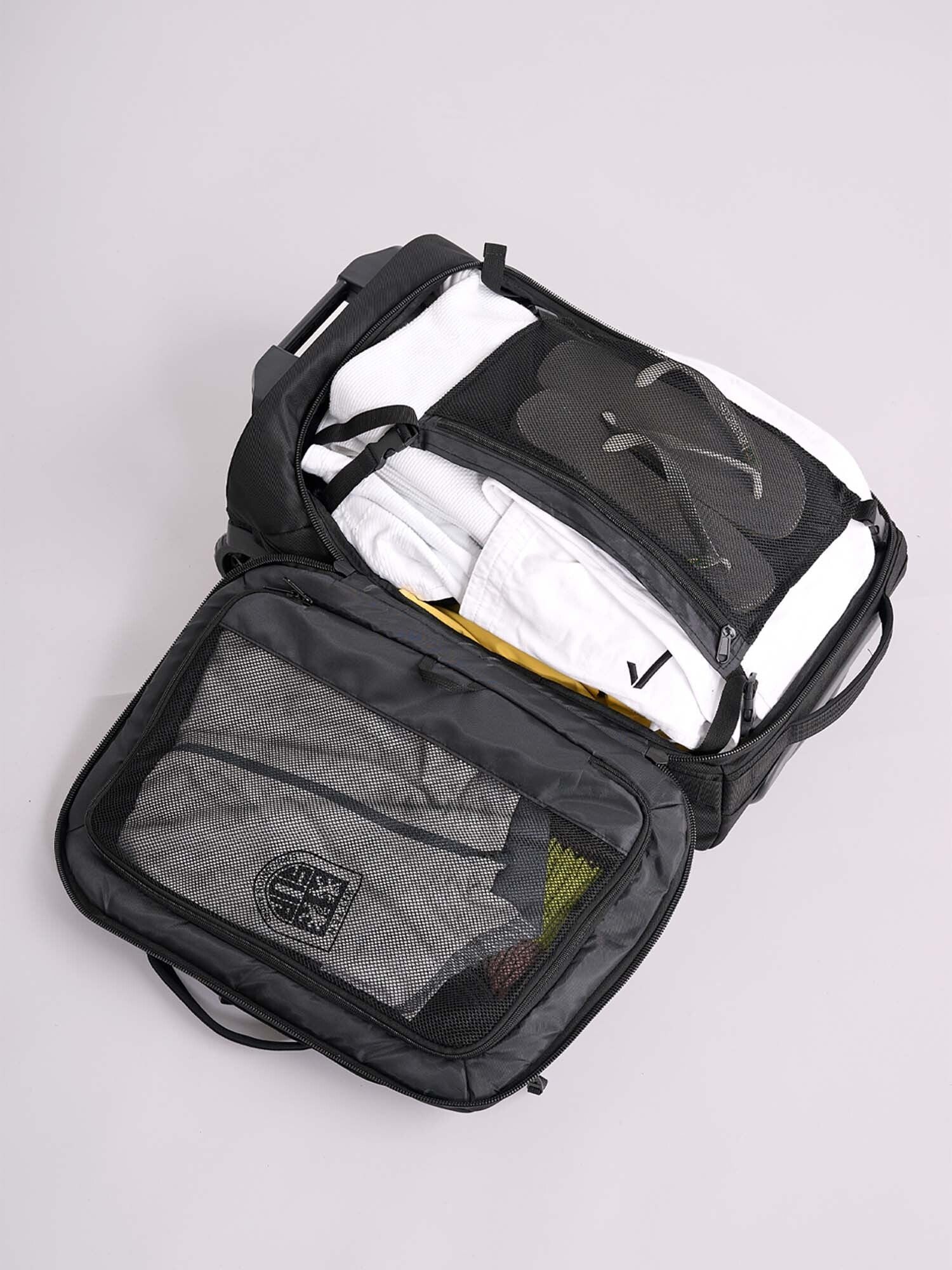 The IPPONGEAR Travel Trolley Black M lies open on a light surface, showing neatly organized contents like folded clothes, black flip-flops, and a mesh divider.