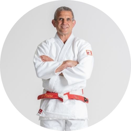 A man wearing a white judo gi and a red and white belt stands with arms crossed, smiling, against a plain light background.