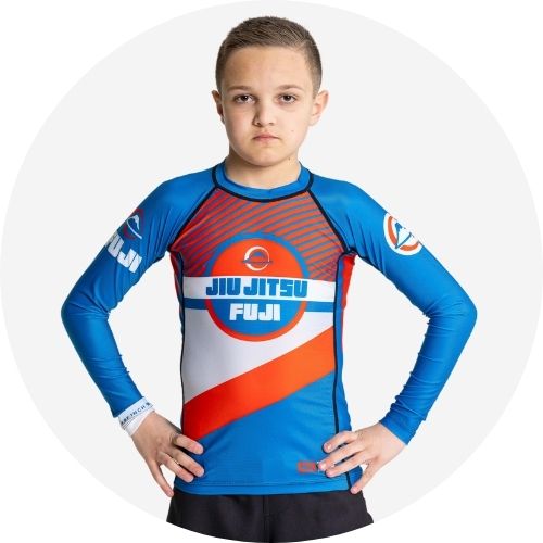 A young boy in a blue and red Fuji Jiu Jitsu rash guard stands with his hands on his hips, looking confidently at the camera against a plain white background.